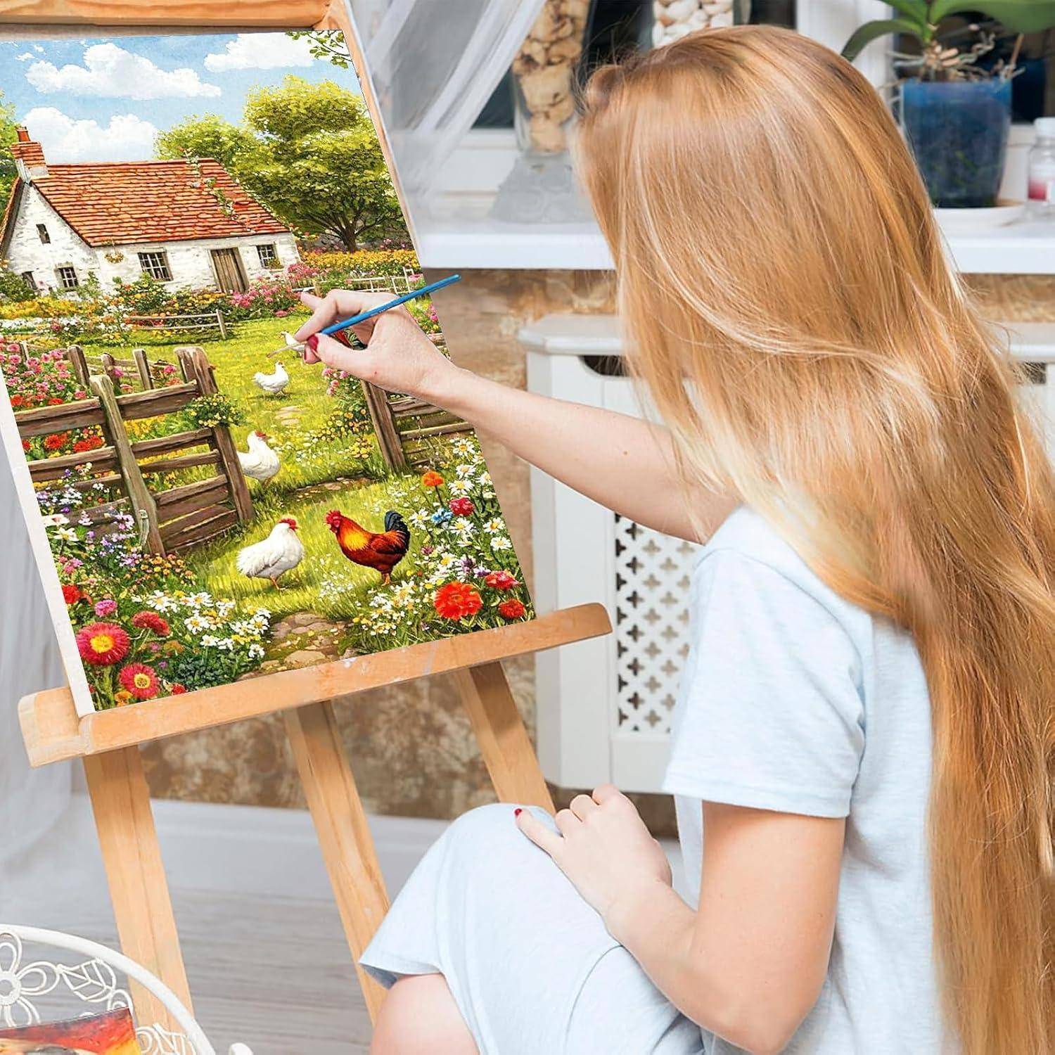 Eine Frau malt eine farbenfrohe Landszene auf einer Staffelei, die einen Bauerngarten mit Blumen, Hühnern und einem Zaun zeigt.