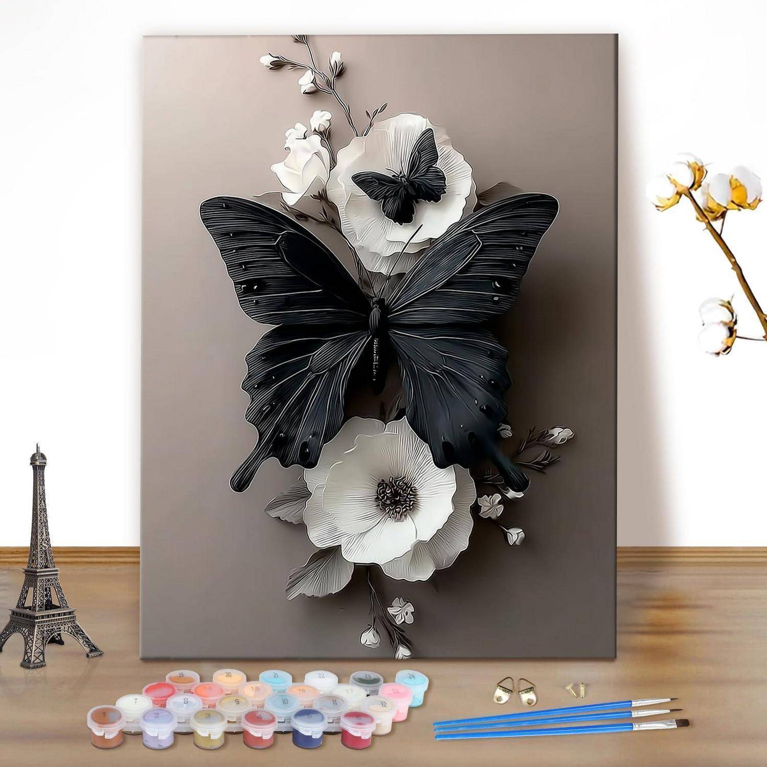 Dekorative Wandkunst in Arbeit: ein großer schwarzer Schmetterling mit weißen Blüten auf einer Leinwand, gezeigt mit Malutensilien auf einem Tisch.