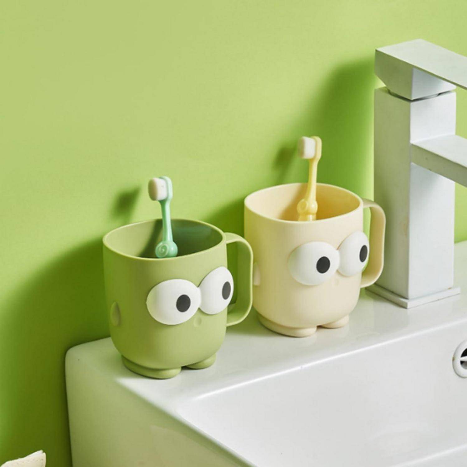 Zwei Zahnputzbecher mit Cartoon-Gesichtern in Grün und Creme stehen auf einem Badezimmerregal mit Zahnbürsten darin.