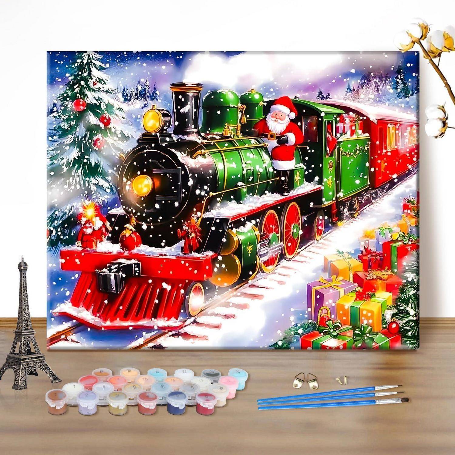 Ein Malen-nach-Zahlen-Set zeigt einen festlichen Weihnachtszug des Weihnachtsmanns im Schnee mit Geschenken, Farbtöpfen, Pinseln und einem kleinen Eiffelturm-Modell auf einem Tisch.
