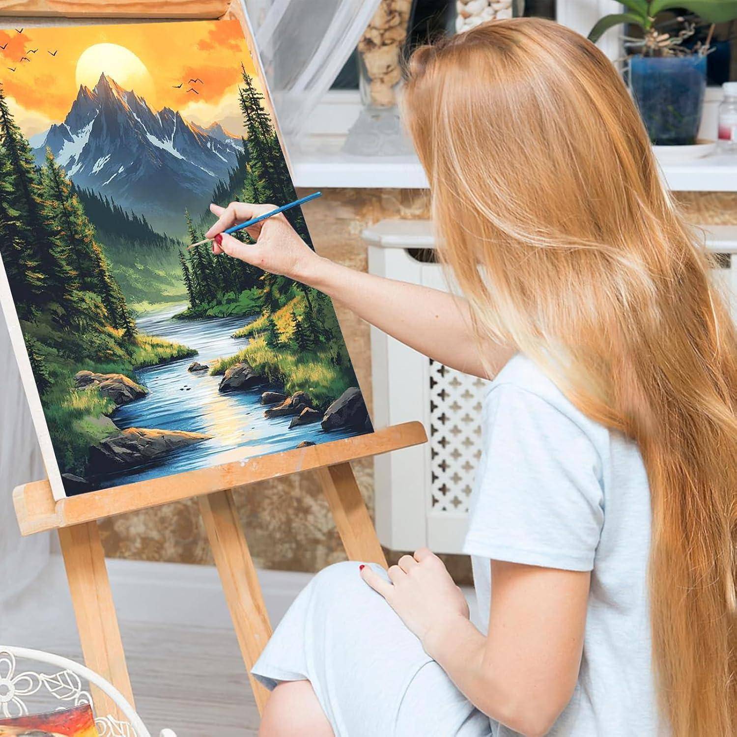 Eine Frau malt eine Berglandschaft auf einer Leinwand an einer Staffelei und fügt Details zu einem Fluss und Bäumen in einem hellen Innenatelier hinzu.