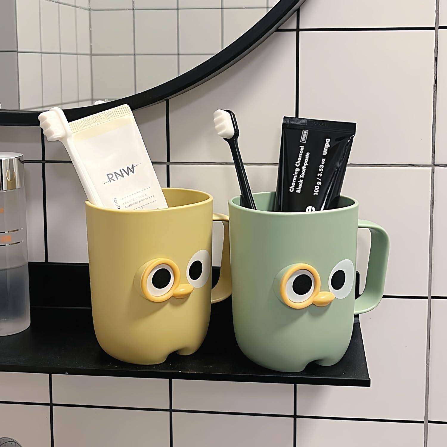 Zwei Cartoon-Gesichts-Becher auf einem Badezimmerregal halten Zahnbürsten und Zahnpastatuben vor einer gekachelten Wand.