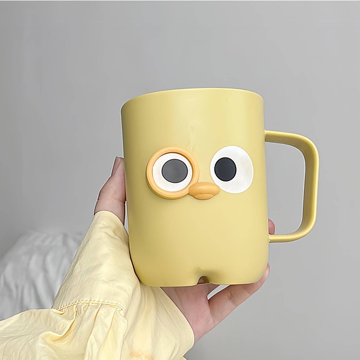Eine gelbe Tasse in Form eines Vogelgesichts mit großen Augen und einer schnabelförmigen Tülle wird in einer Hand gegen einen einfachen Hintergrund gehalten.