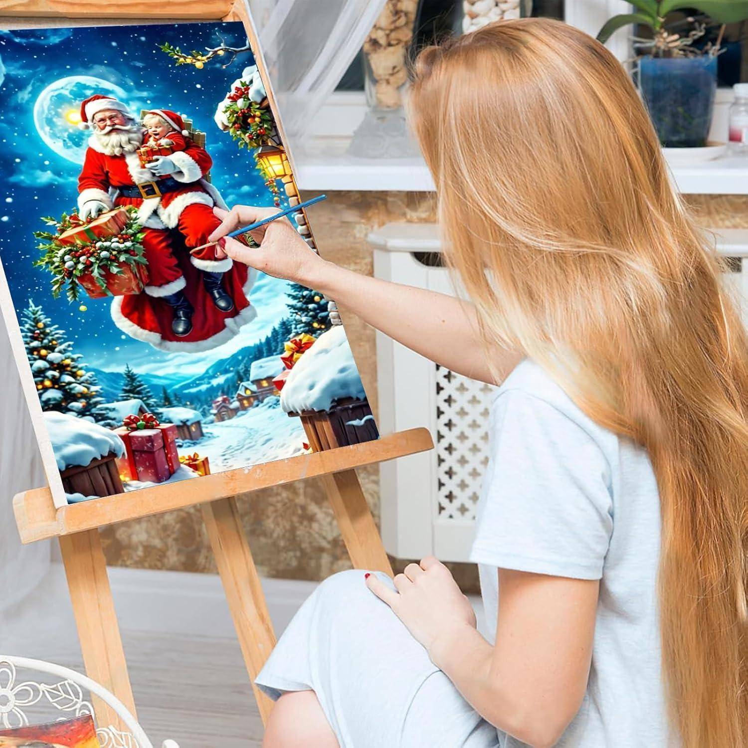 Eine Frau malt eine festliche Weihnachtsszene mit dem Weihnachtsmann und einem Kind in einem roten Schlitten auf einer Staffelei in einem hellen Zimmer.