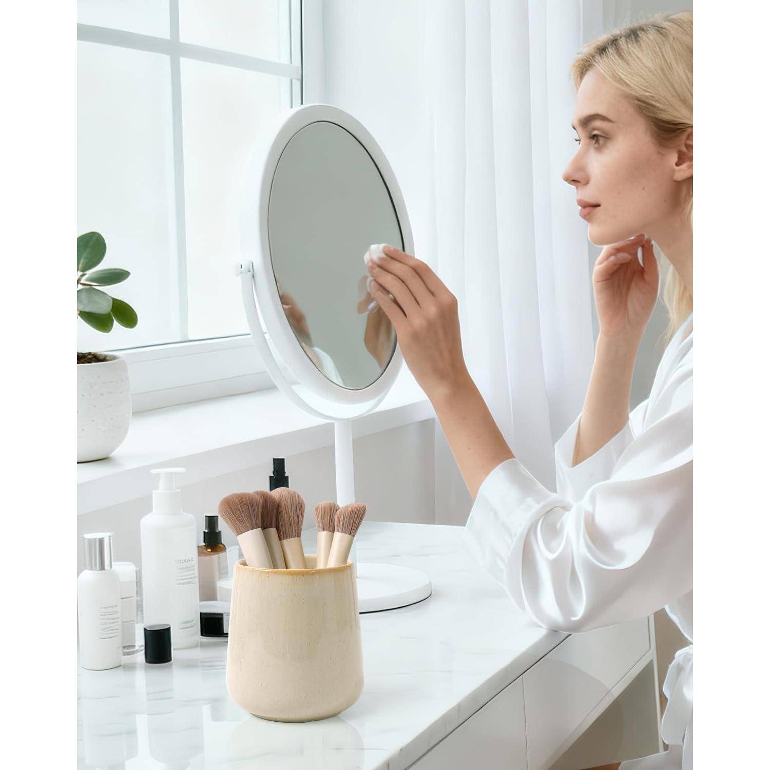 Frau in einem weißen Bademantel, die mit einem Wattestäbchen Make-up aufträgt, während sie in einen Kosmetikspiegel neben Hautpflegeflaschen und Pinseln neben einem Fenster schaut.