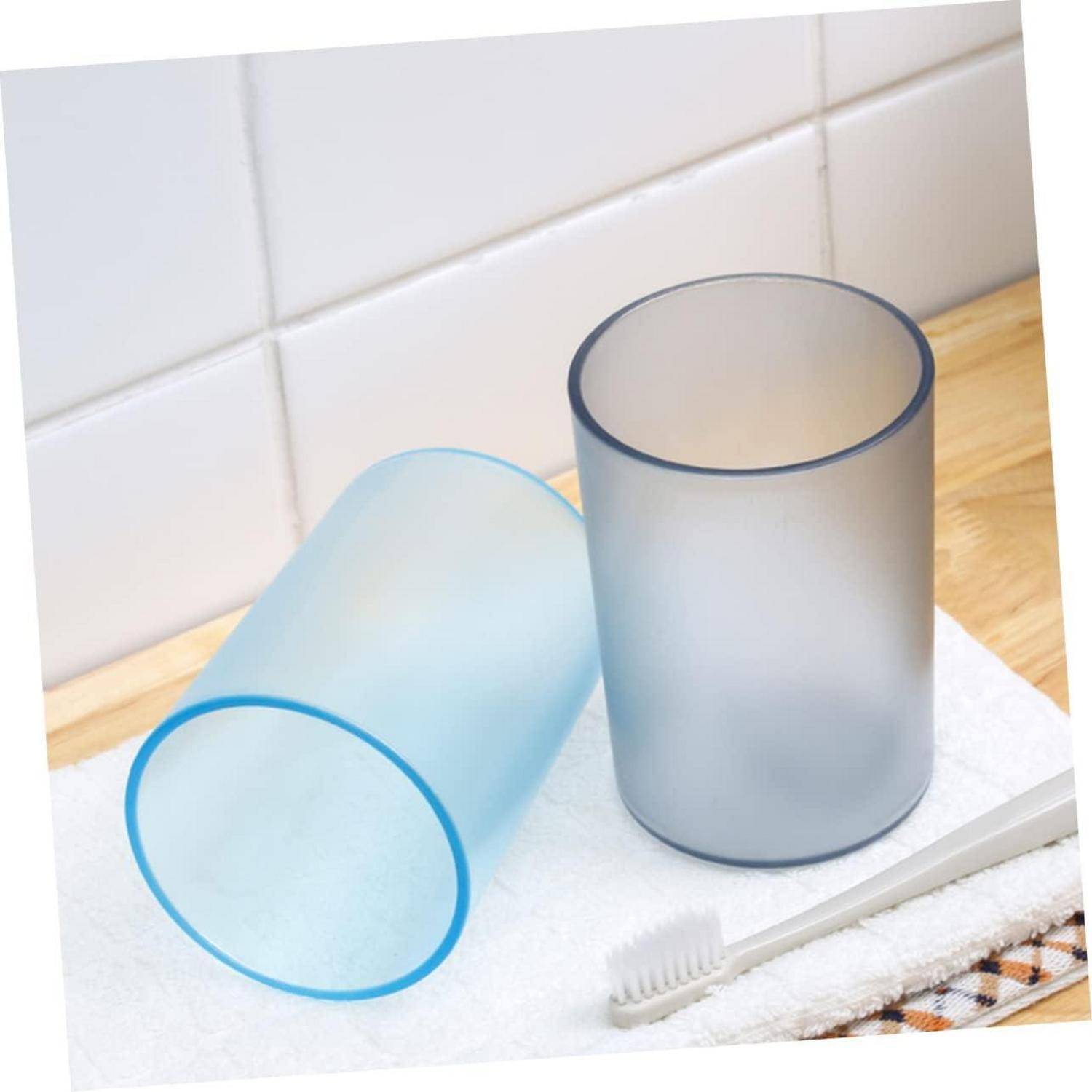 Zwei durchscheinende mattierte Kunststoffbecher, einer aufrecht und einer auf der Seite liegend, auf einem Badezimmerregal mit einer Zahnbürste und einem Handtuch.