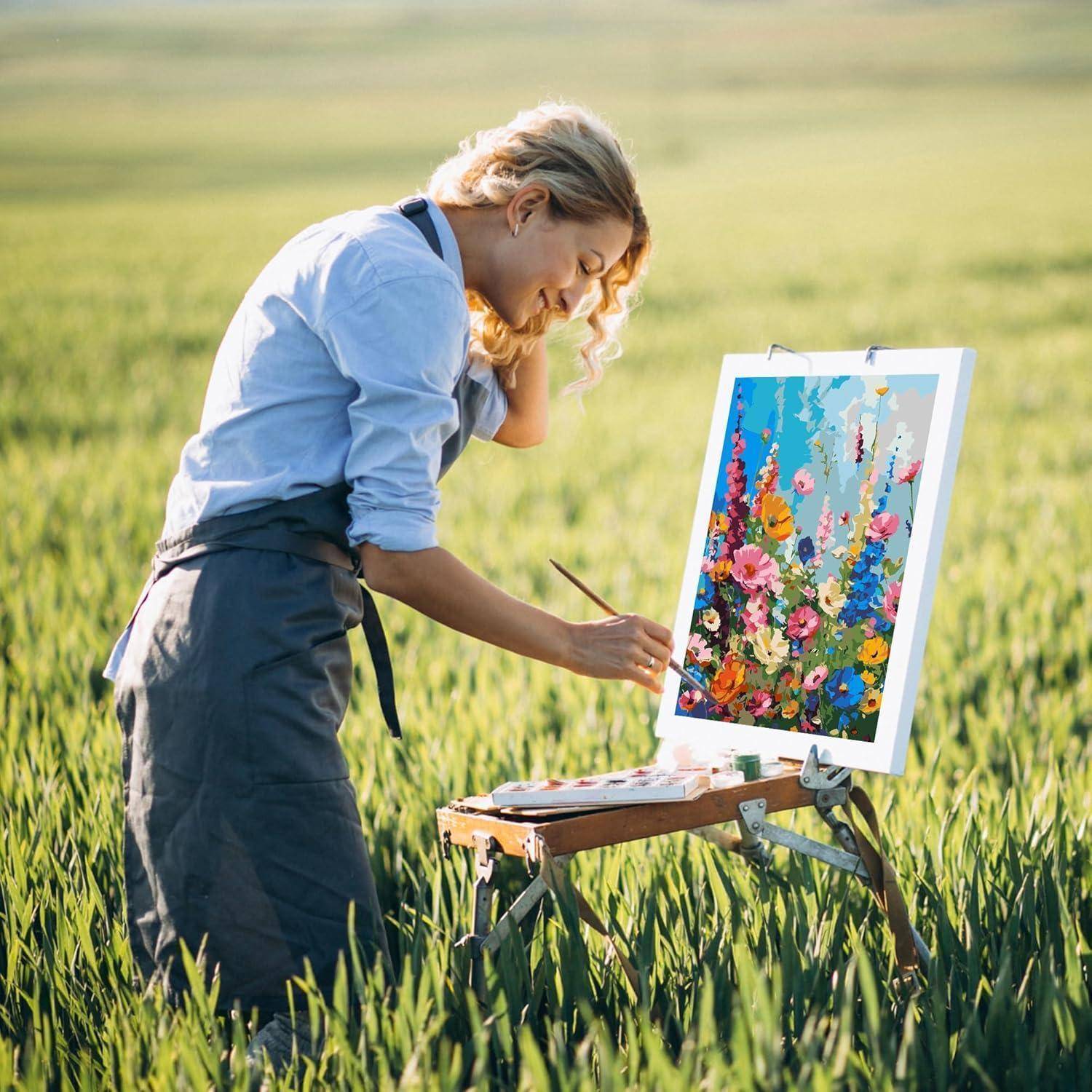 Eine Frau malt eine bunte Blumenszene auf einer Staffelei in einem Grasfeld im Freien.