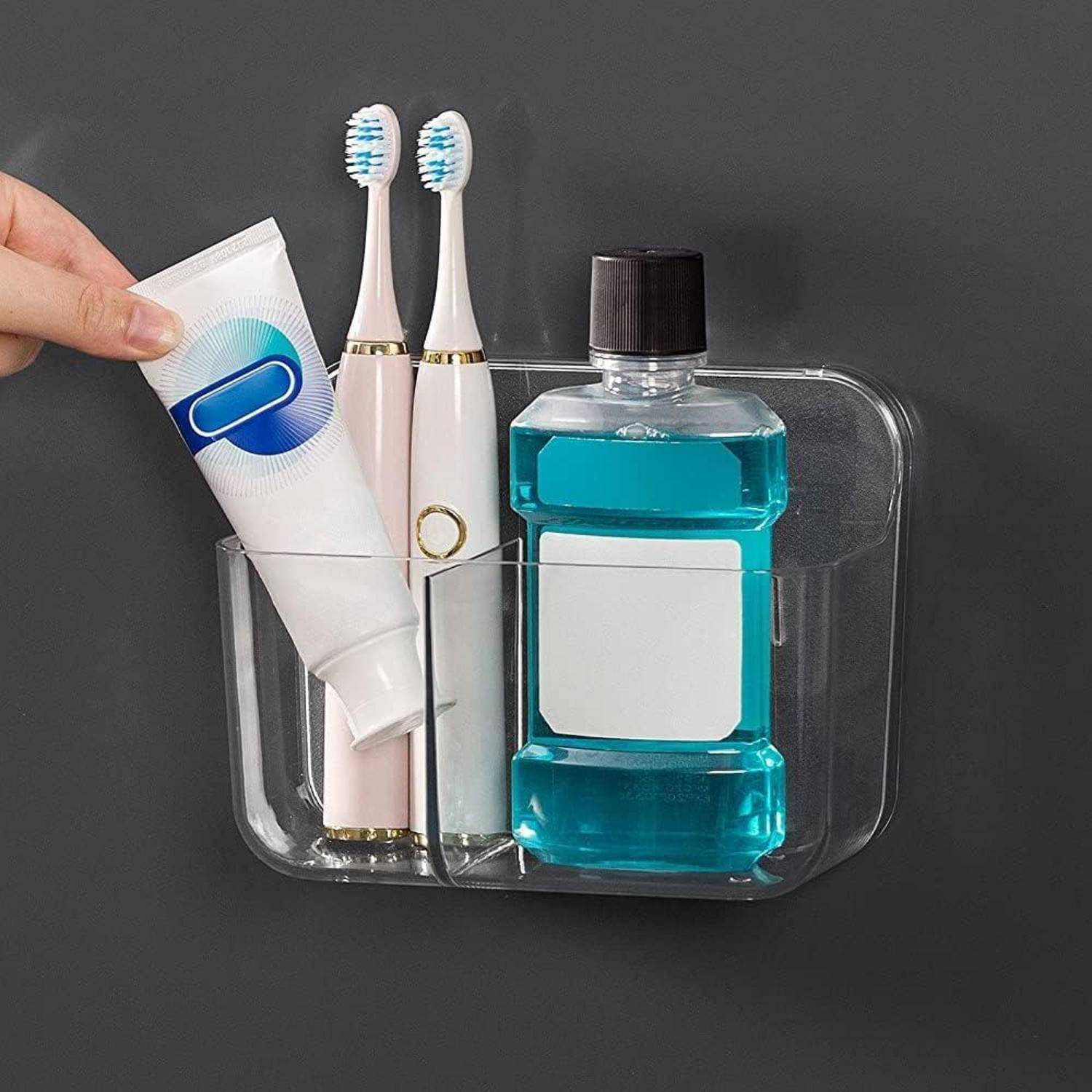 Wandmontierter transparenter Badorganizer mit zwei elektrischen Zahnbürsten, Zahnpasta und einer Mundspülungsflasche, während eine Hand die Zahnpastatube platziert.