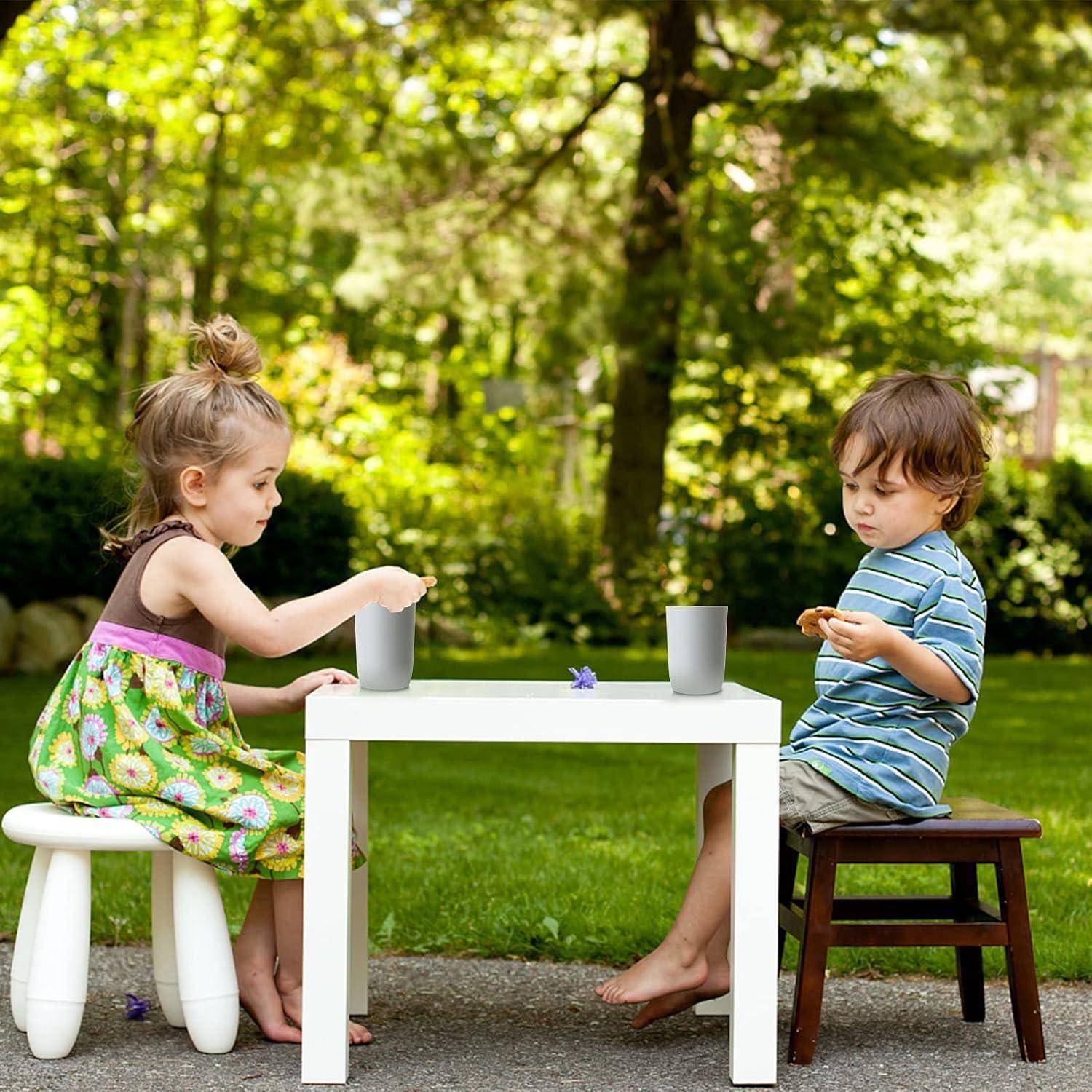 Zwei kleine Kinder sitzen an einem kleinen weißen Tisch im Freien und spielen mit Bechern in einem Garten.