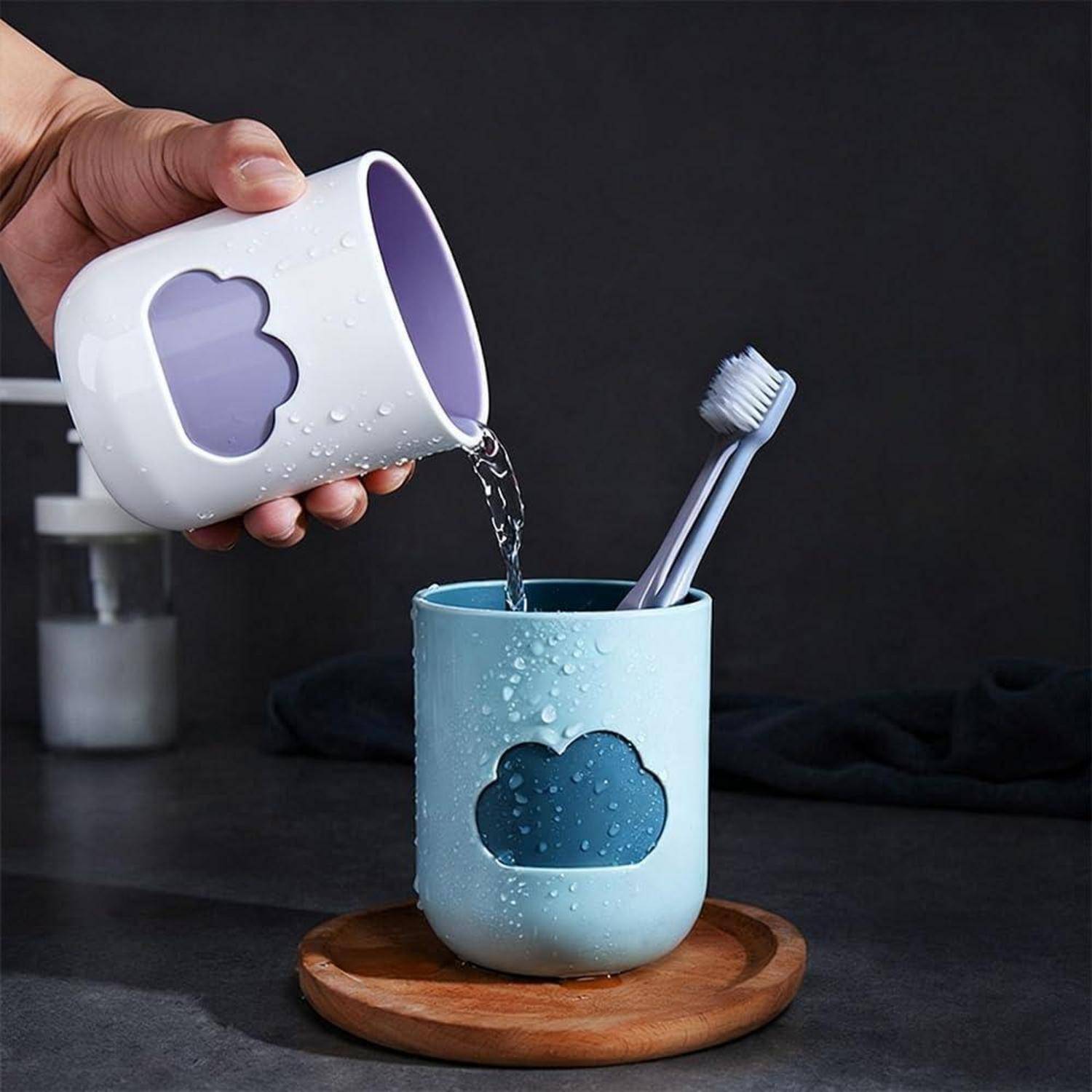 Eine Hand gießt Wasser aus einer weißen Tasse in einen blauen Zahnbürstenbecher, der zwei Zahnbürsten auf einem hölzernen Tablett hält.