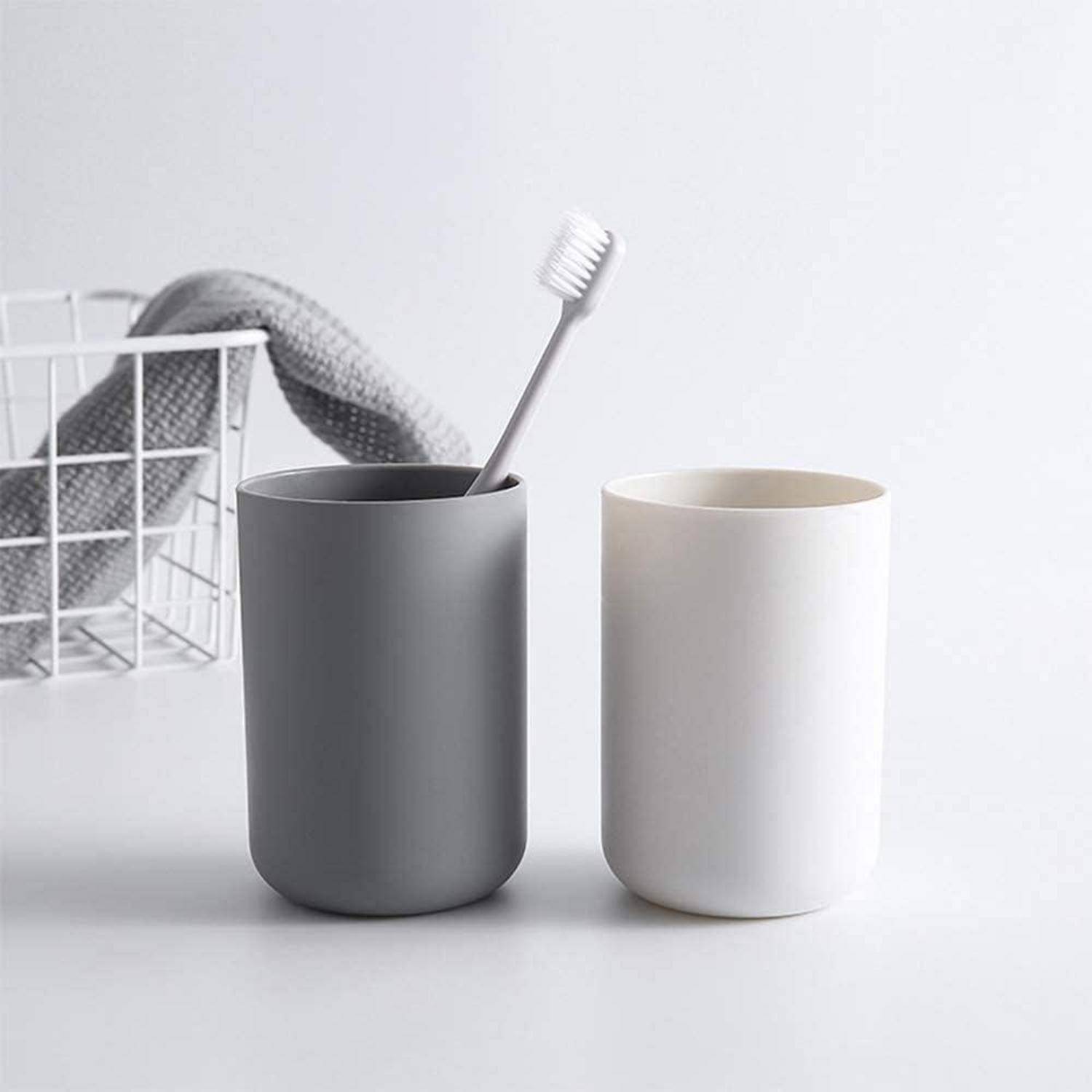 Zwei minimalistische Badezimmertassen, eine grau mit einer Zahnbürste und eine weiß, auf einer weißen Oberfläche neben einem Drahtkorb mit einem Tuch.