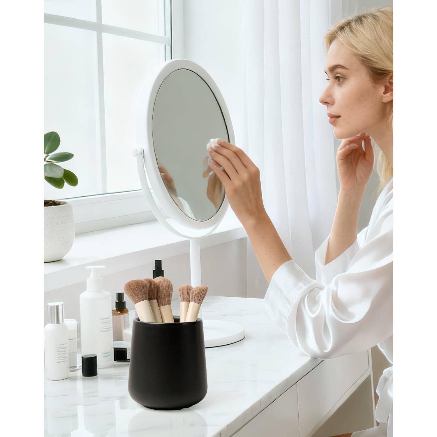 Frau in einem weißen Bademantel trägt Hautpflege- oder Make-up-Produkte auf, während sie in einen Schminkspiegel schaut, neben einem Tisch mit Kosmetikprodukten und Pinseln.