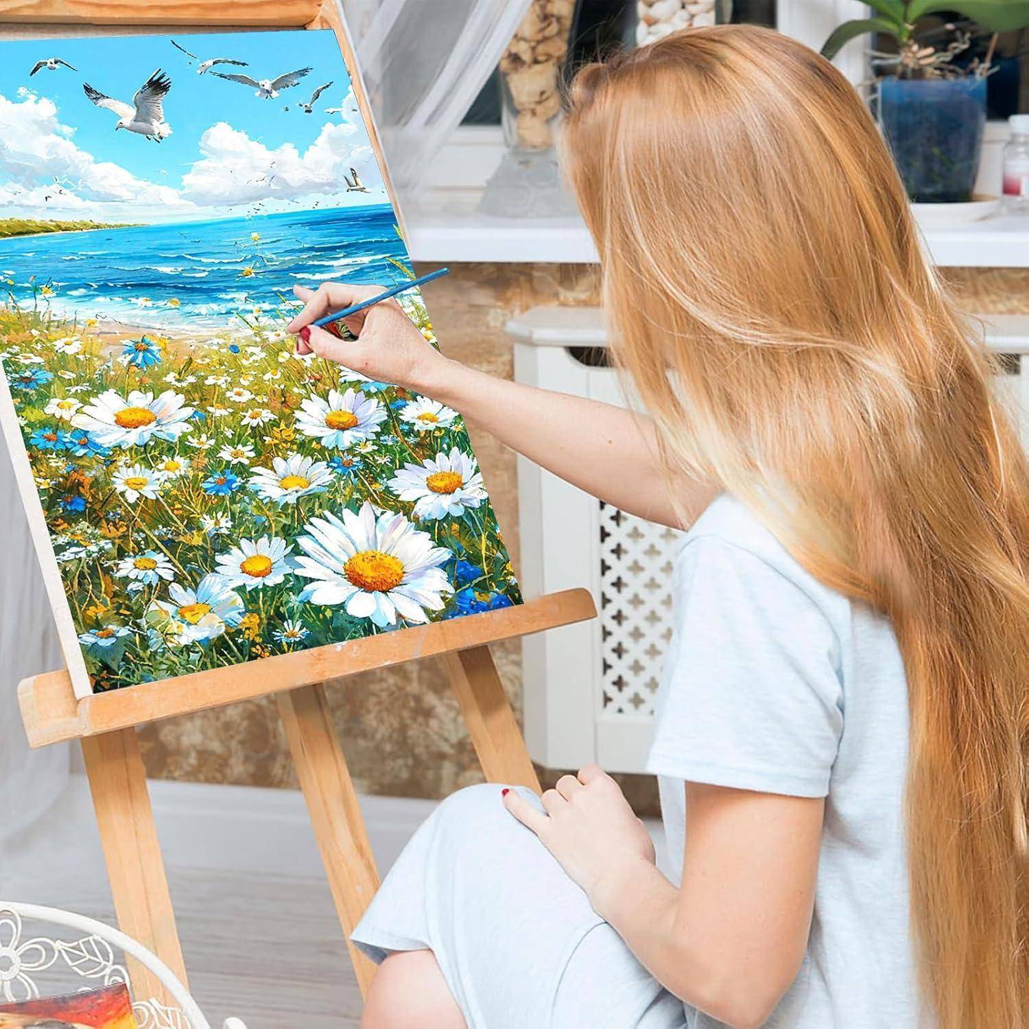 Junge Frau malt eine Strandlandschaft mit Gänseblümchen und fliegenden Möwen auf einer Staffelei im Innenraum.