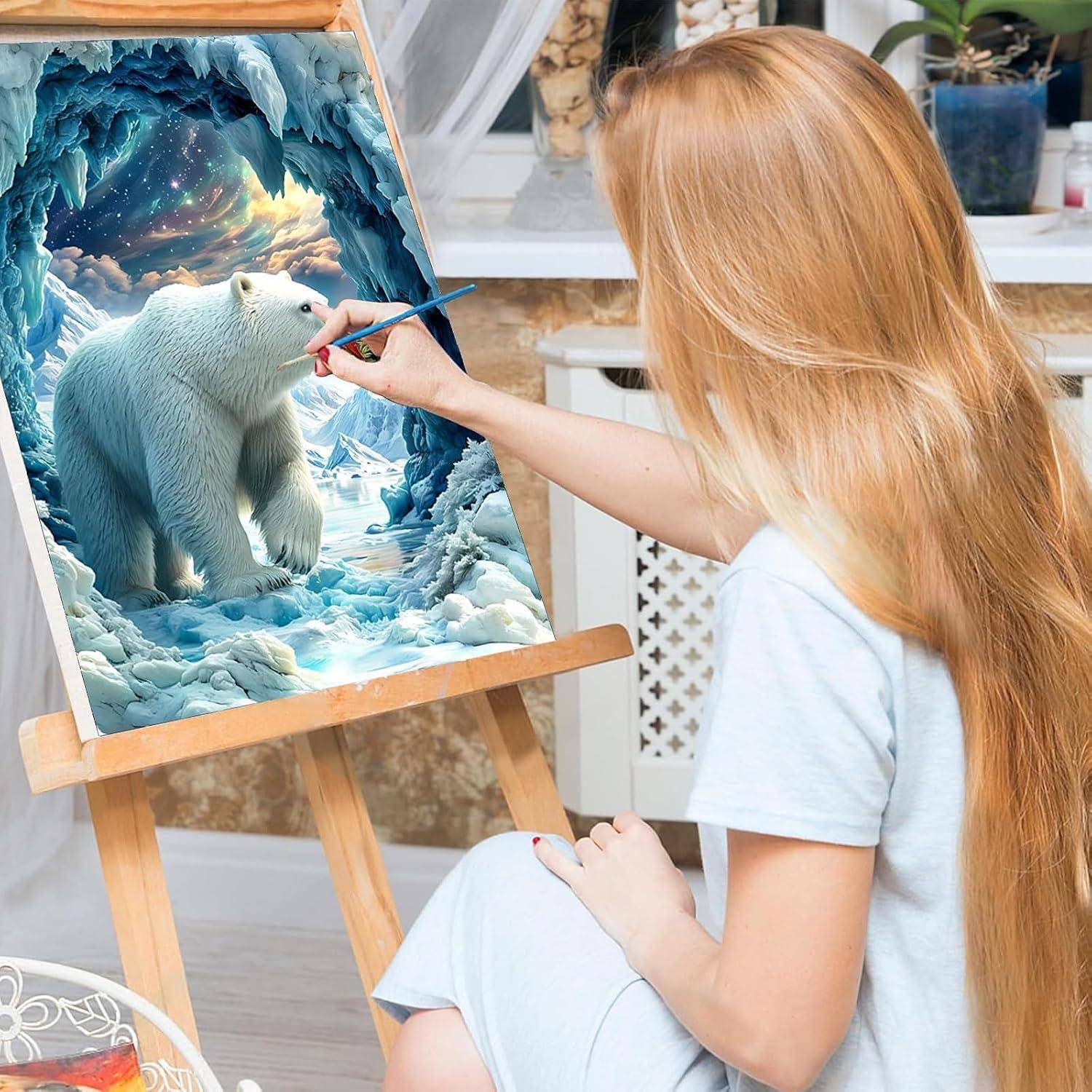 Blonde Frau malt einen Eisbären in einer eisigen Höhlenszene auf einer Staffelei, wobei das fertige Kunstwerk einen Sternenhimmel und blaue Eisformationen zeigt.