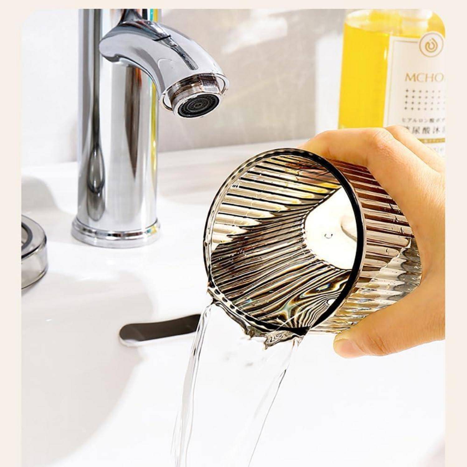 Hand gießt Wasser aus einem gerippten Metallbecher in ein Badezimmerbecken unter einem Chromhahn.