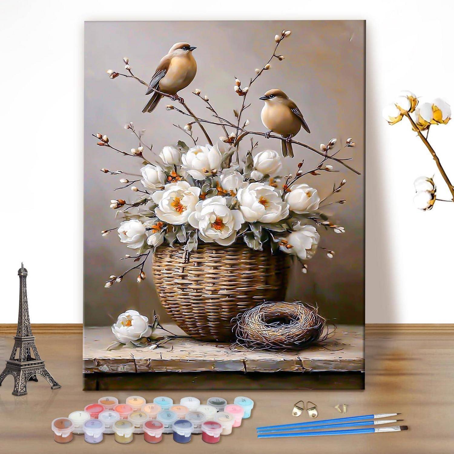 Kunstmaterialien auf einem Tisch vor einer Leinwandmalerei mit einem Weidenkorb voller weißer Blumen, zwei Vögeln und einem Nest; Eiffelturm-Dekoration in der Nähe.