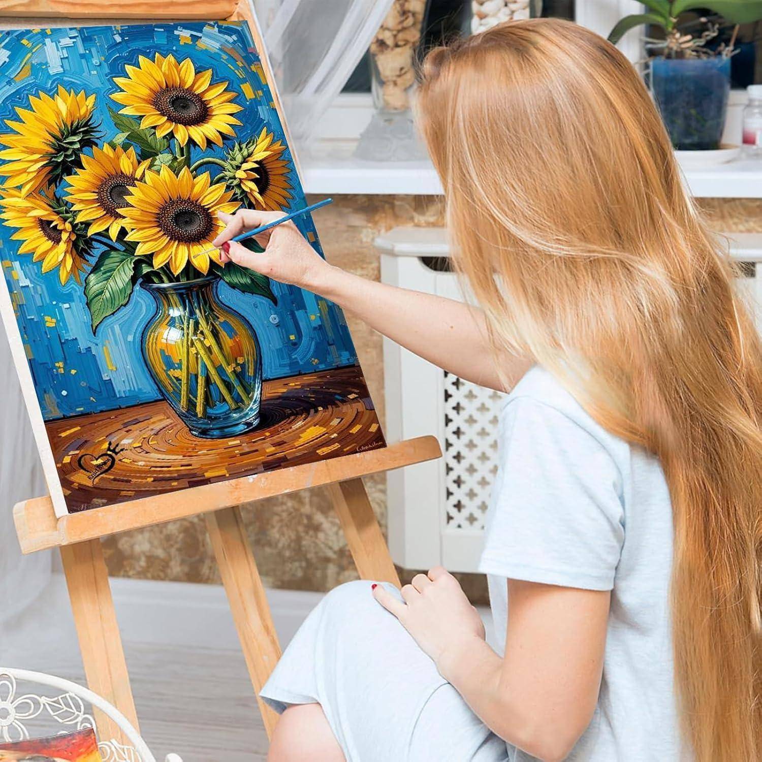 Eine blonde Frau malt ein Sonnenblumen-Stillleben auf einer Staffelei und fügt Details zu einer Vase mit Blumen in einem hellen Innenraum-Studio hinzu.