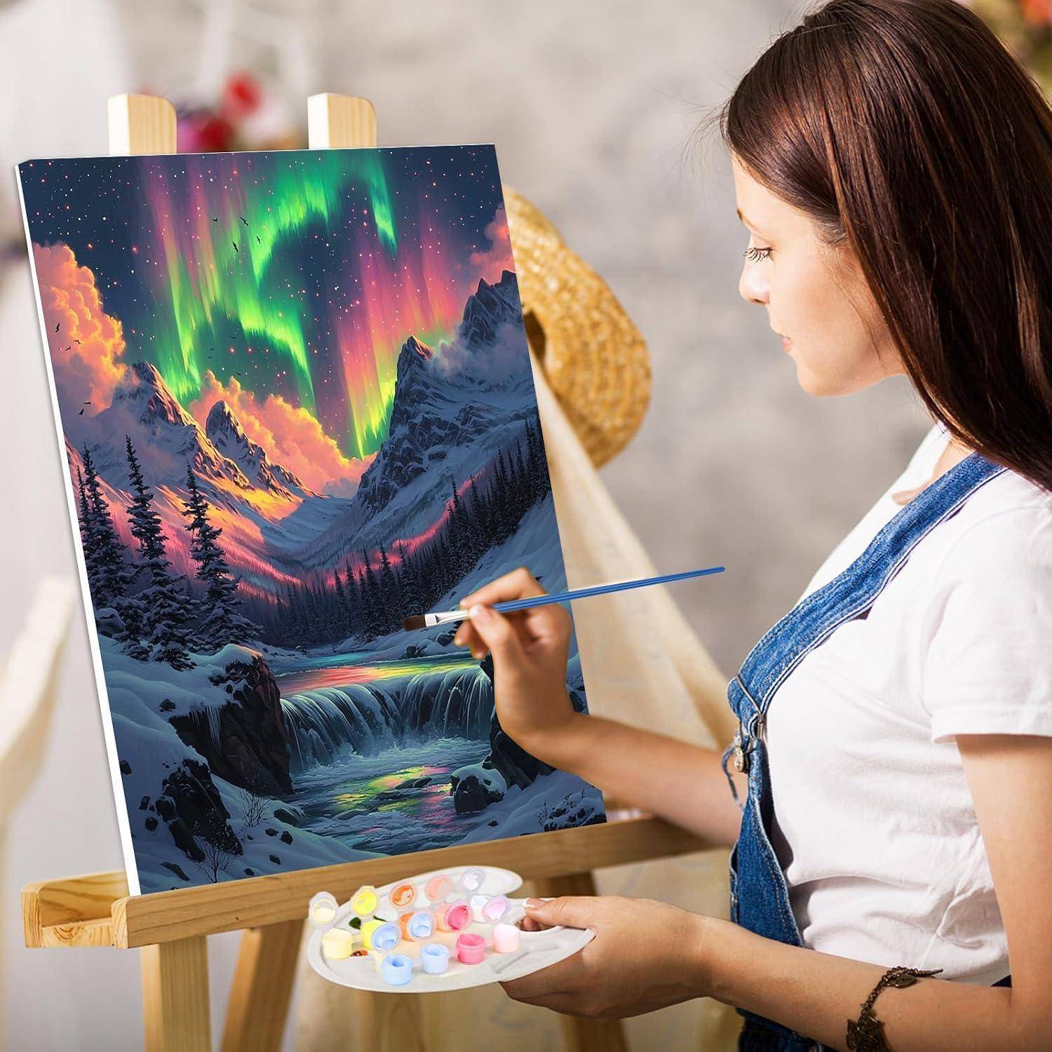 Eine Frau malt eine Winterlandschaft auf einer Staffelei und fügt Details zu einem farbenfrohen Nordlicht über verschneiten Bergen und einem Wasserfall hinzu.