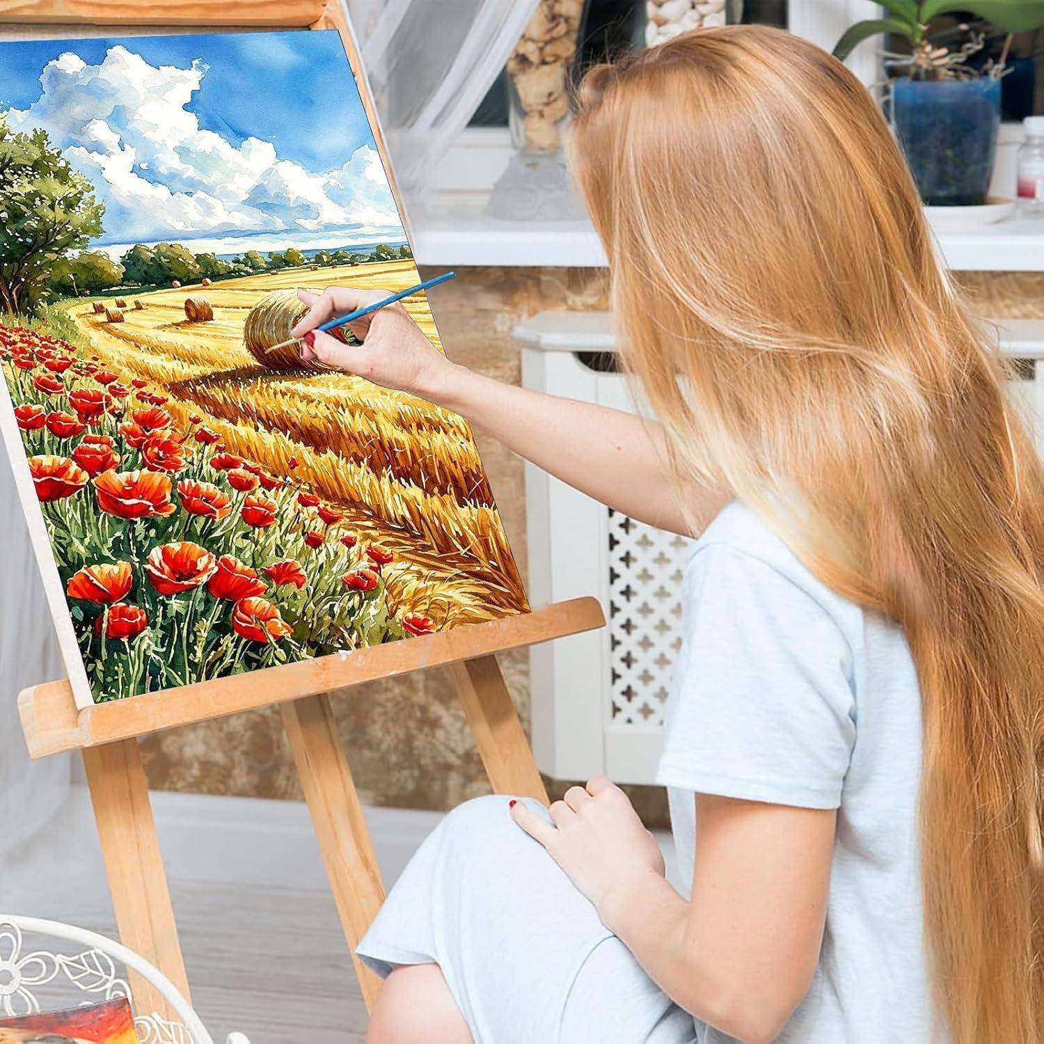 Frau malt eine farbenfrohe Feldlandschaft auf einer Staffelei im Innenraum, mit roten Blumen, Heuballen und einem bewölkten blauen Himmel.