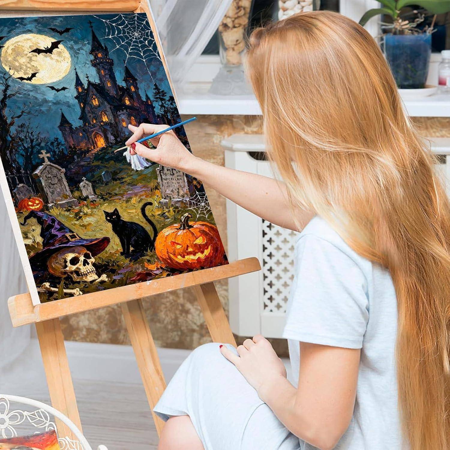 Eine Frau malt eine Halloween-Szene auf einer Staffelei, die ein Spukhaus, einen Mond, Spinnennetze, eine schwarze Katze, einen Schädel und Kürbisse zeigt.