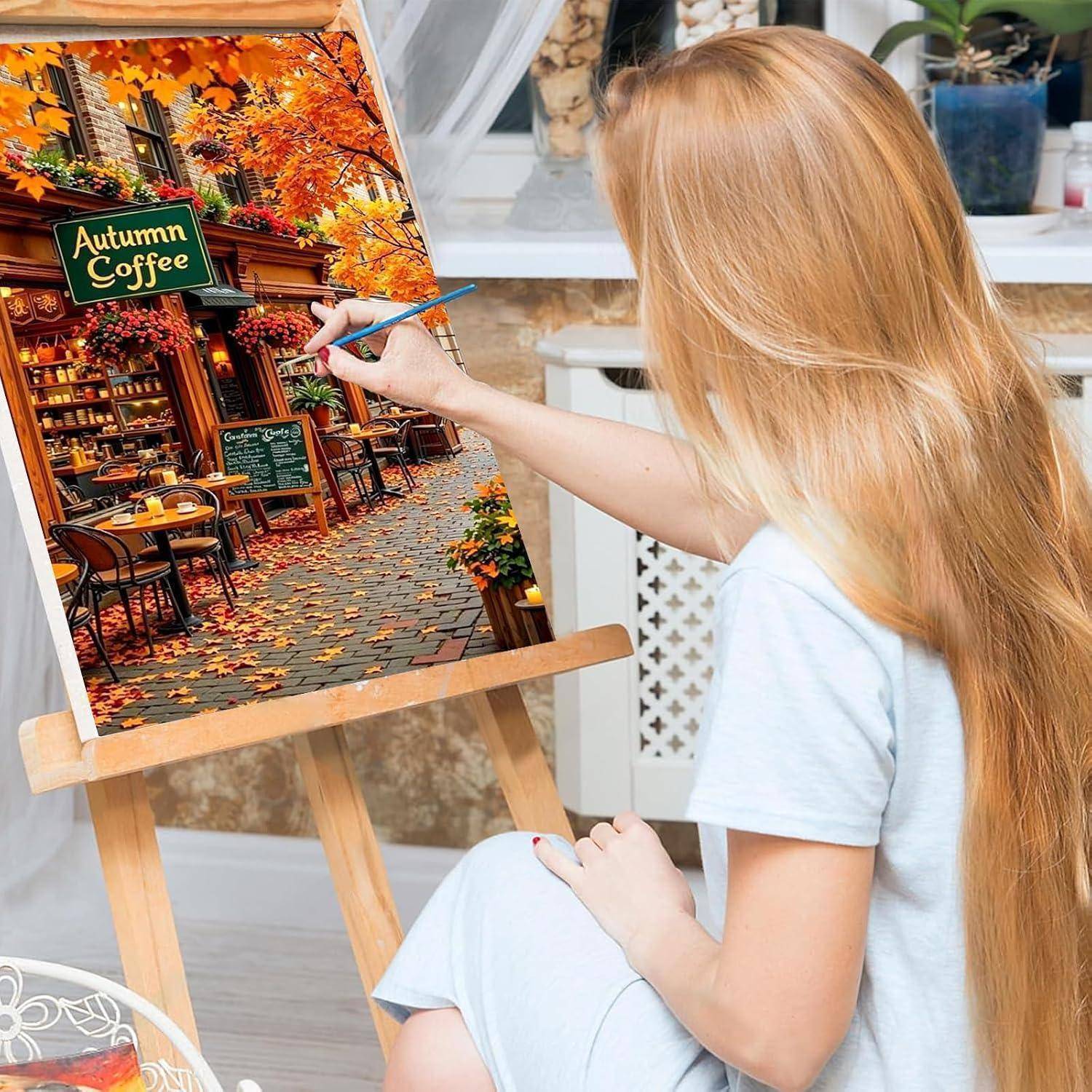 Eine blonde Frau malt eine Herbstcafé-Szene auf eine Leinwand, mit orangefarbenen Blättern, Außentischen und einem Schild mit der Aufschrift 
