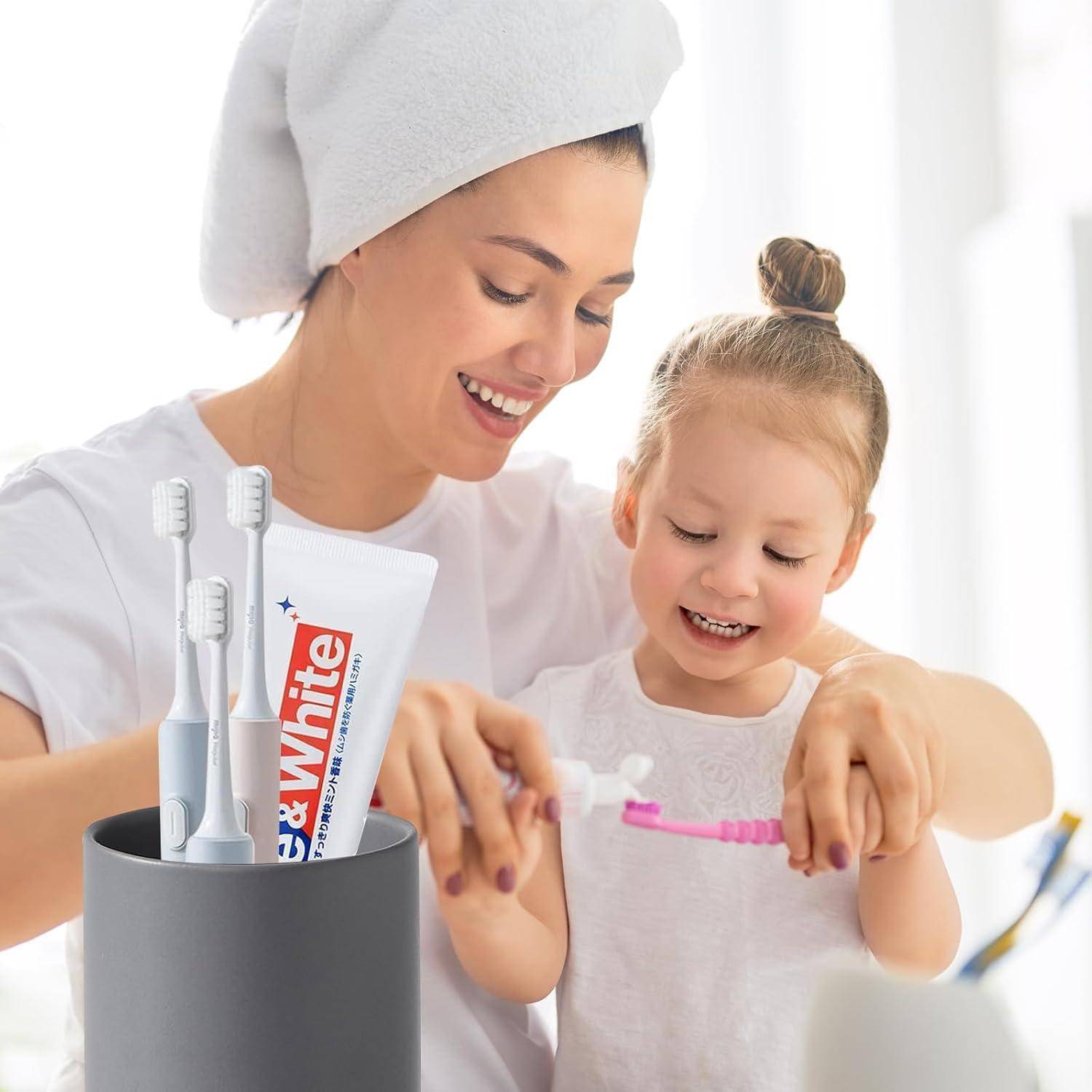 Mutter und kleine Tochter lächeln sich an, während sie zusammen die Zähne putzen, mit elektrischen Zahnbürsten und Zahnpasta im Badezimmer.