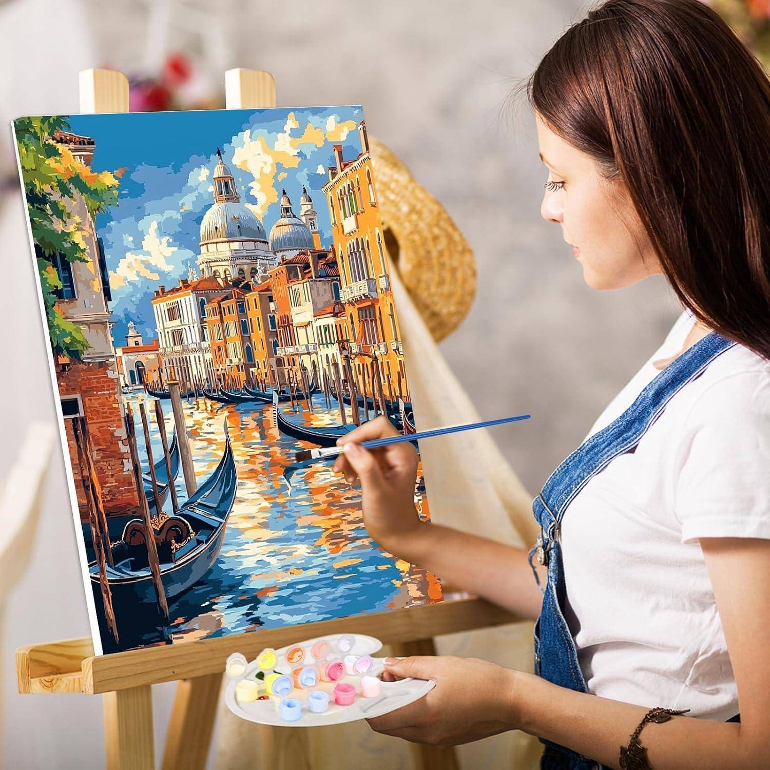 Eine Frau malt eine farbenfrohe Kanalszenerie einer Stadt auf eine Leinwand an einer Staffelei und hält dabei eine Palette und einen Pinsel in den Händen.
