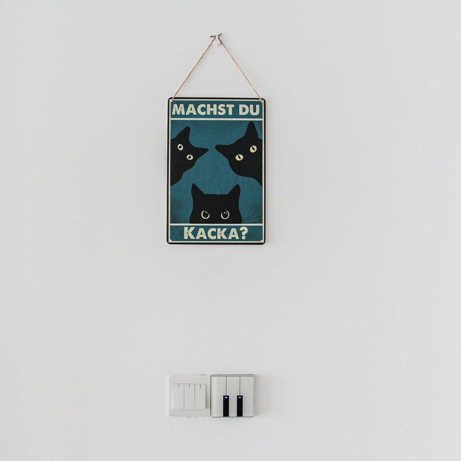 Ein kleines Hängeschild mit drei schwarzen Katzengesi­chtern und deutschem Text „MACHST DU KACKA?