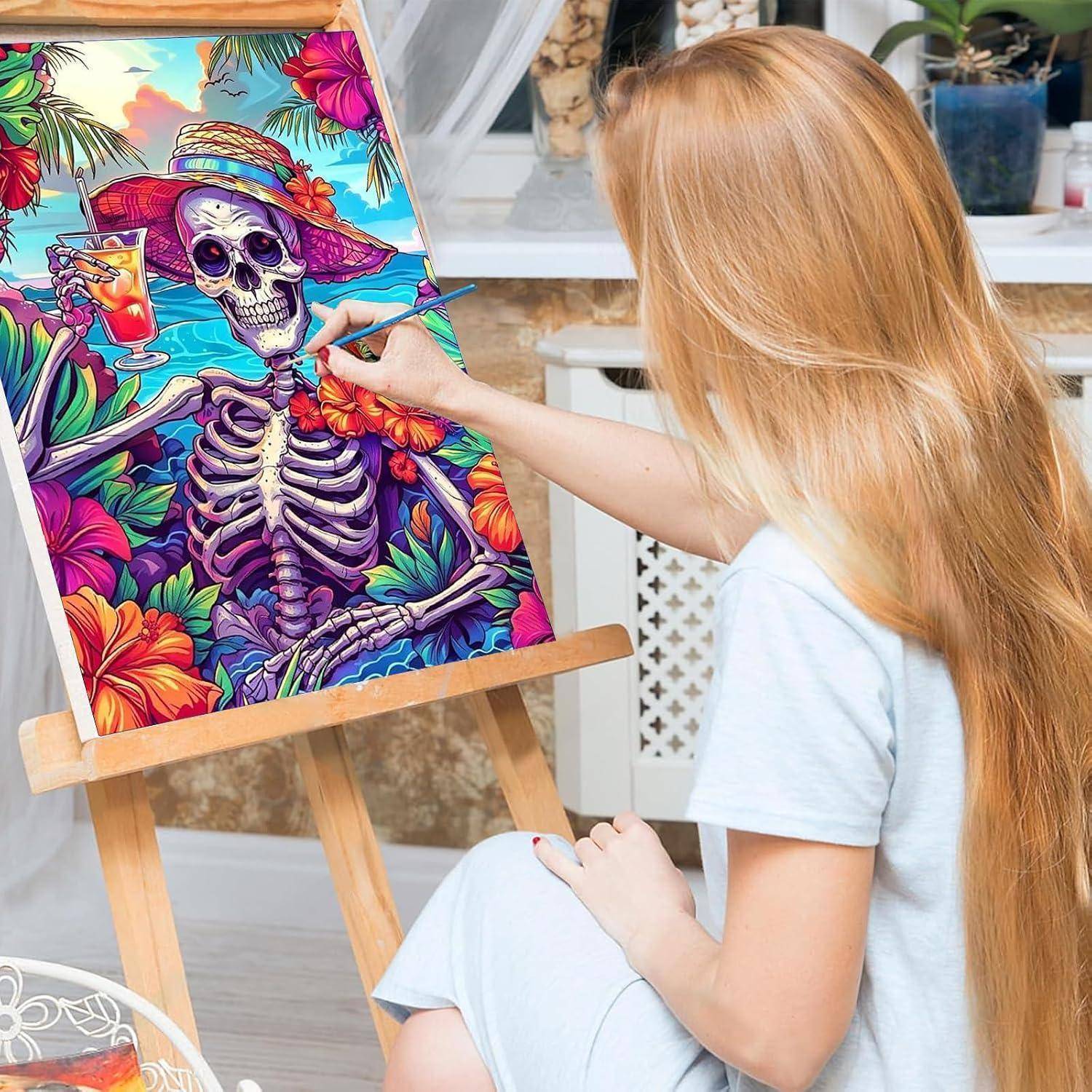 Eine Frau malt eine bunte, totenkopfthematisierte tropische Szene auf eine Leinwand auf einer Staffelei und hält einen Pinsel in einem hellen Studio.