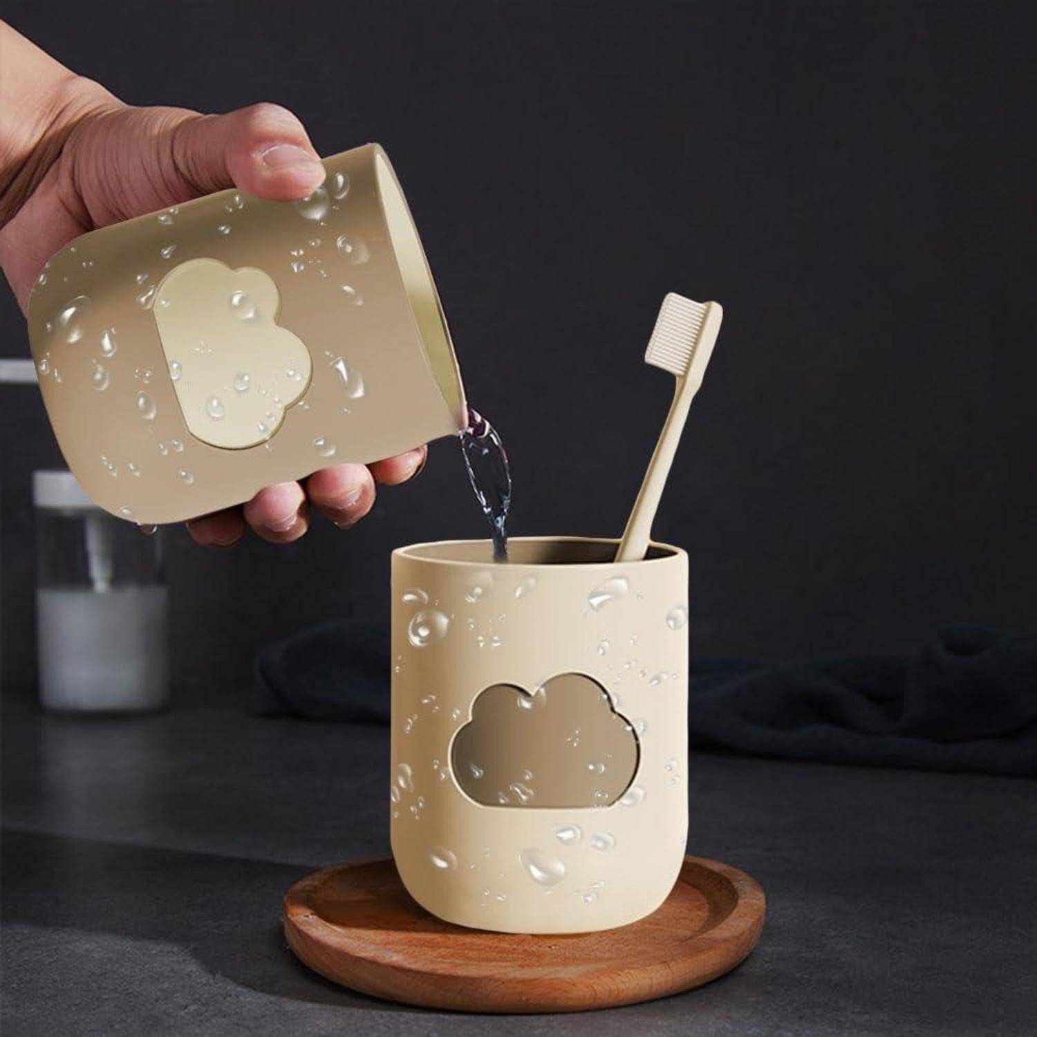 Eine Hand gießt Wasser aus einer passenden beigen Tasse in einen wolkenförmigen Zahnbürstenhalter, der eine Zahnbürste auf einem hölzernen Tablett hält.