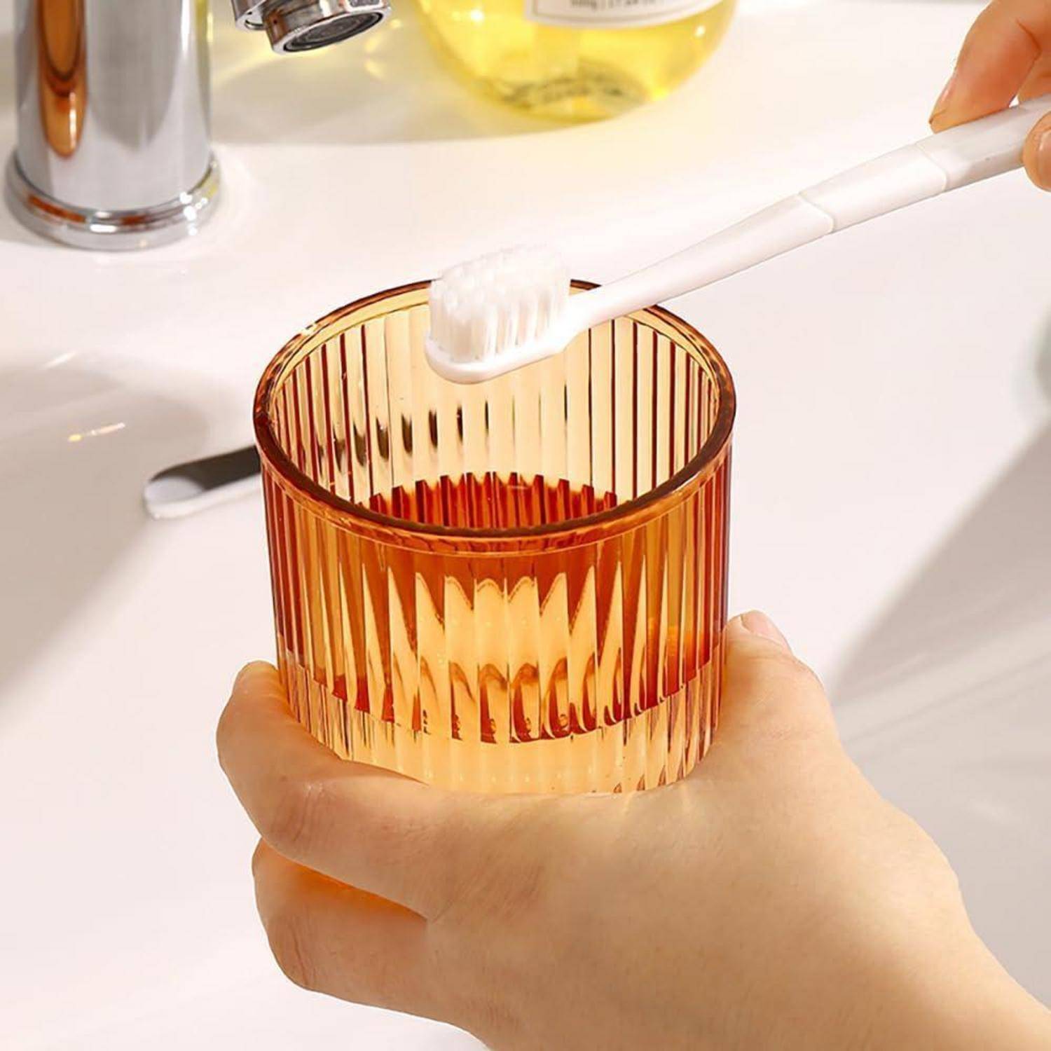 Eine Hand hält ein Bernsteinglas, während eine Zahnbürste darüber unter fließendem Wasser an einem Badezimmerbecken gespült wird.