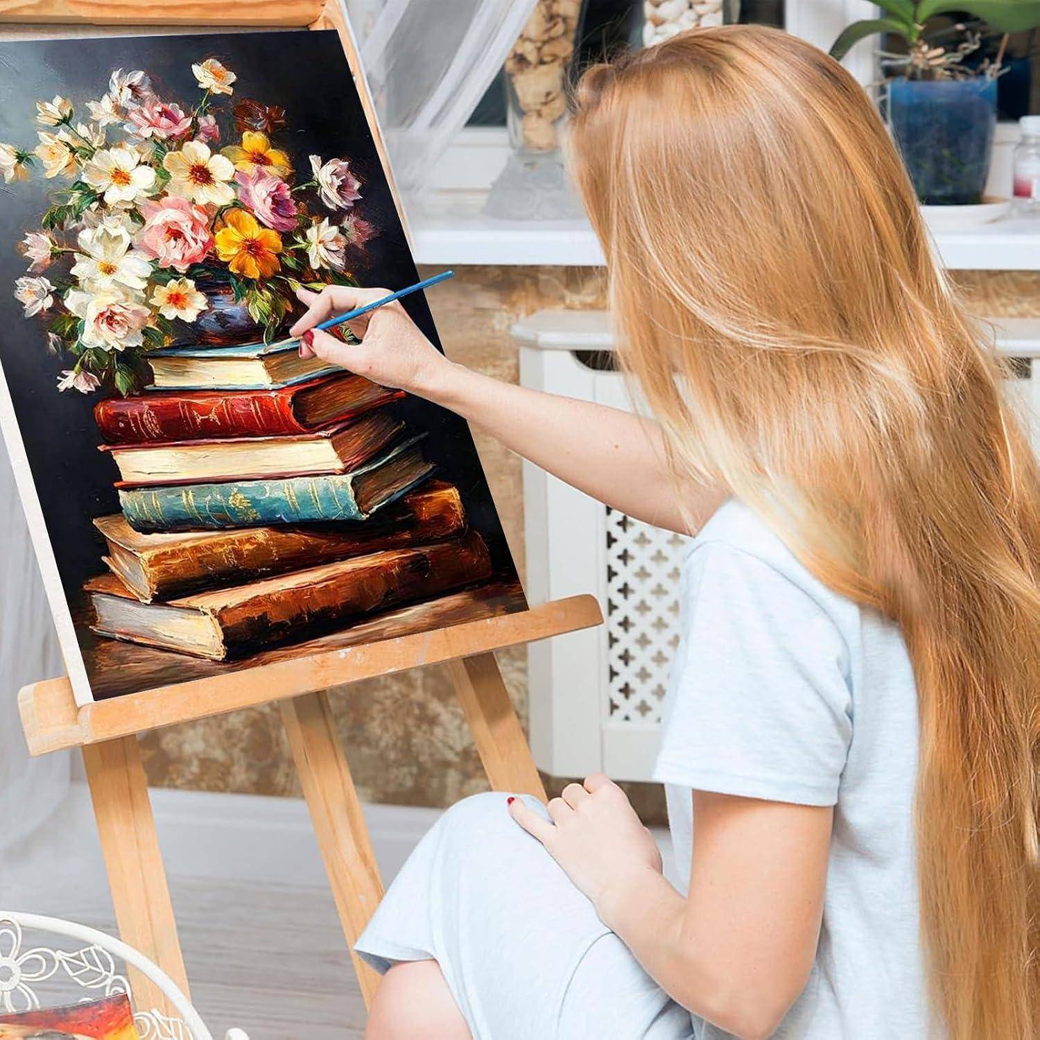 Eine Frau mit langen blonden Haaren malt Blumen in einer Vase auf einer Staffelei, wobei ein Stapel alter Bücher das Stillleben bildet.