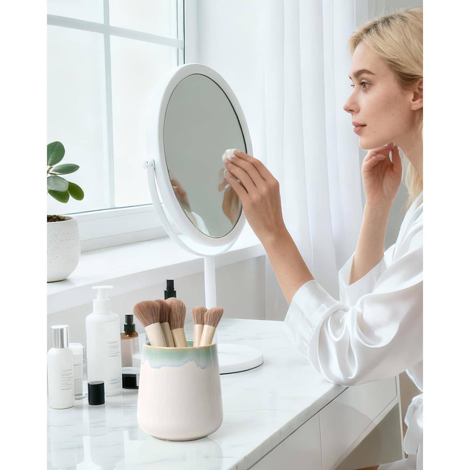 Frau in einem weißen Bademantel trägt Hautpflege auf, während sie in einen Kosmetikspiegel schaut, neben Makeup-Pinseln und Schönheitsprodukten neben einem hellen Fenster.