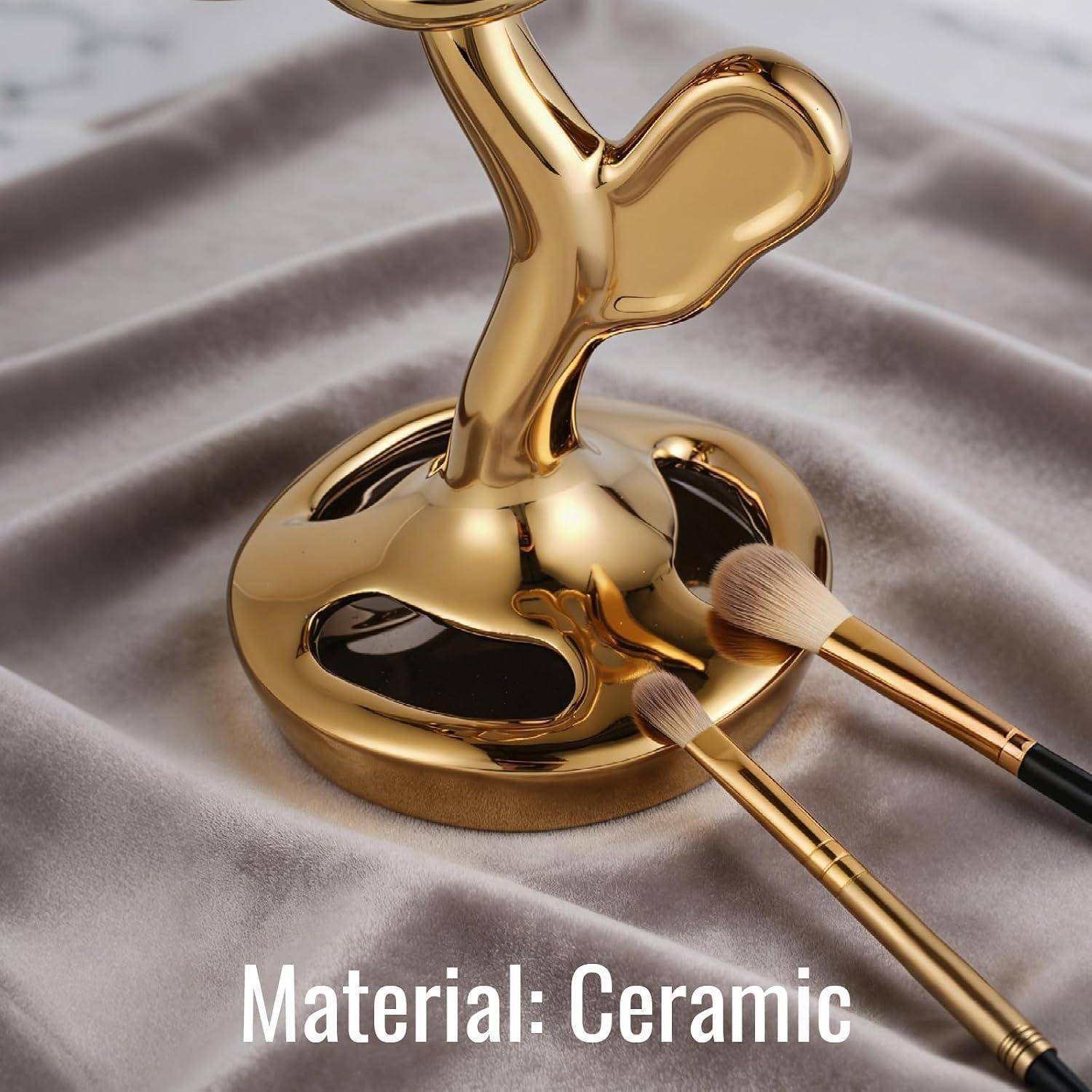 Goldener Keramik-Kosmetikpinselhalter mit zwei Make-up-Pinseln auf einem drapierten Stoffhintergrund; Text lautet „Material: Keramik