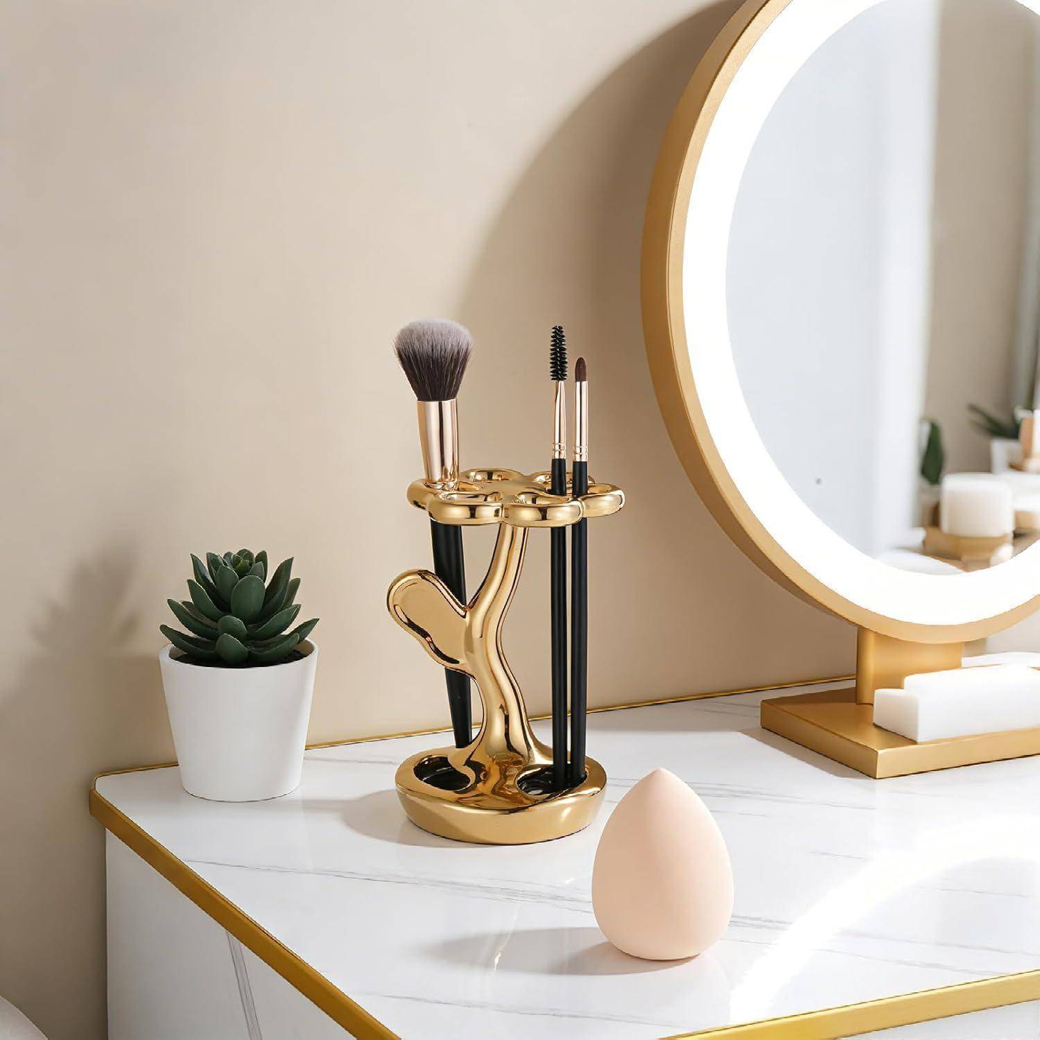 Halter für Goldmakeup-Pinsel und Spiegel auf einem Waschtisch mit weißer Marmoroberfläche, eine kleine Sukkulente und ein Make-up-Schwamm.