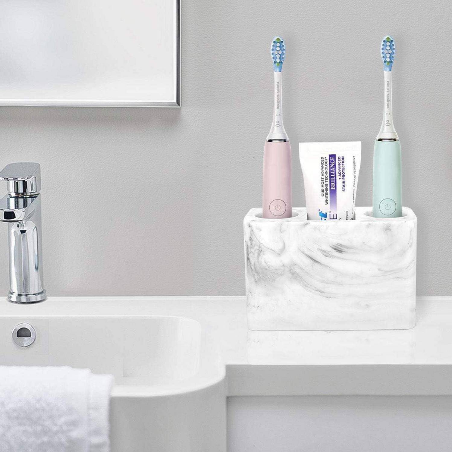 Zwei elektrische Zahnbürsten und Zahnpasta in einem Marmor-Badezimmerhalter neben einem Waschbecken und einem Wasserhahn.