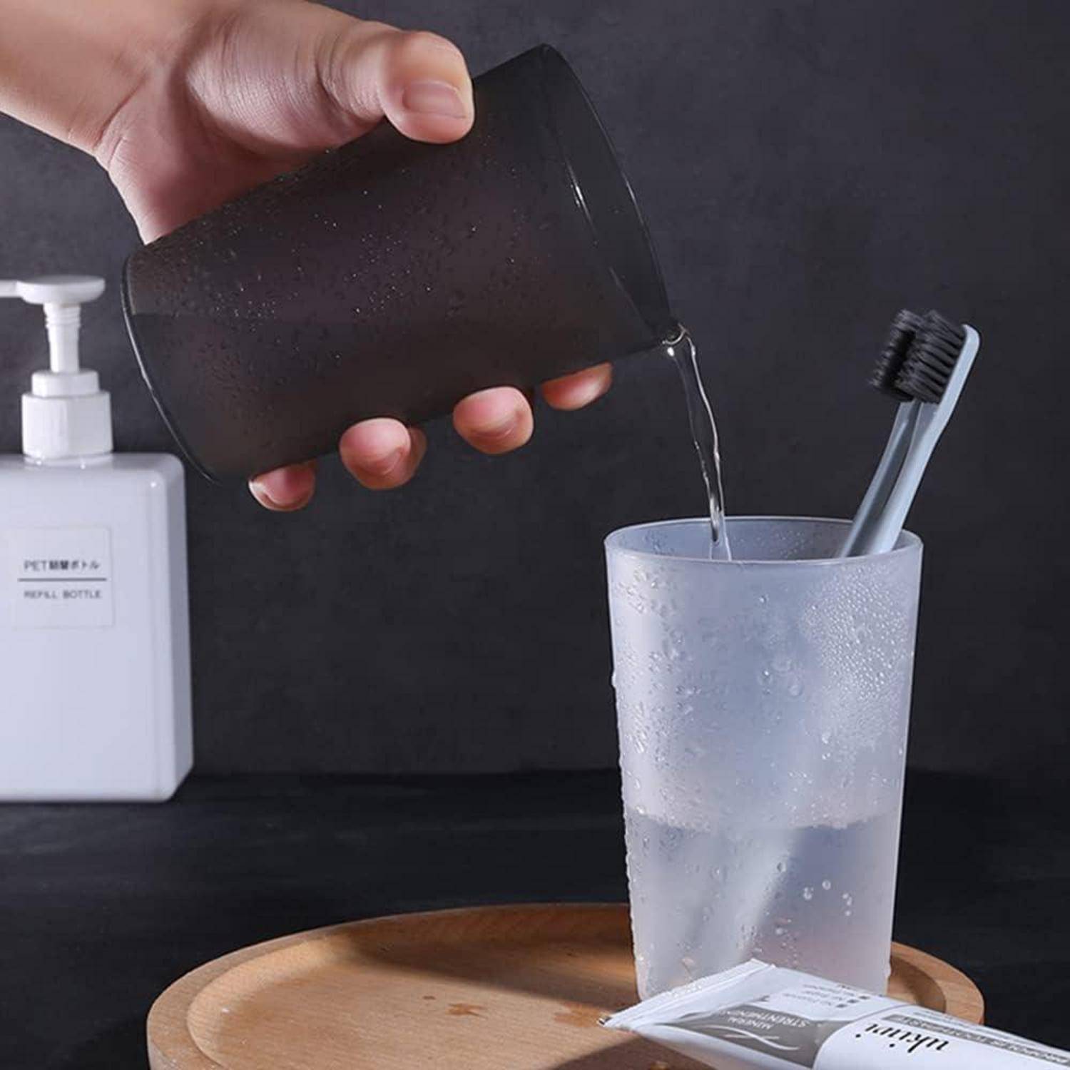 Eine Hand gießt Flüssigkeit aus einer schwarzen Tasse in einen mattierten Badezimmerwasserbecher, der eine Zahnbürste auf einem Tablett hält.