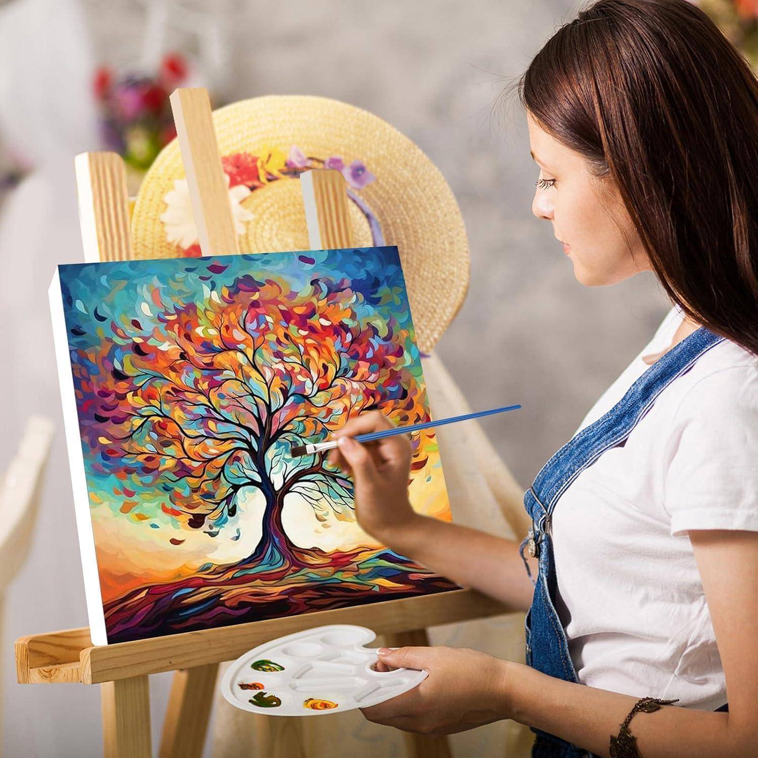 Eine junge Frau malt einen farbenfrohen abstrakten Baum auf einer Leinwand an einer Staffelei und hält eine Palette und einen Pinsel in einem Atelier.