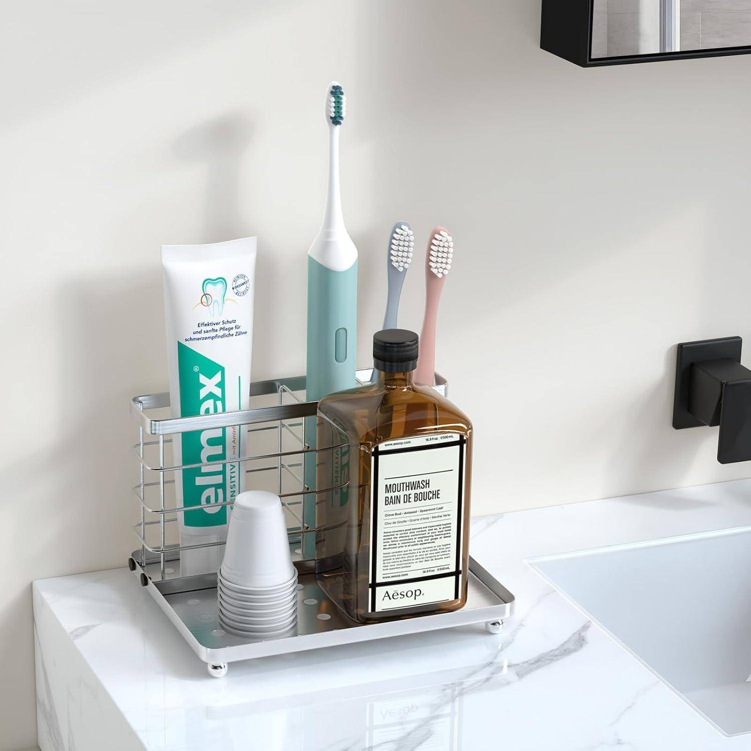 Badezimmer-Arbeitsflächenorganizer mit Zahnpasta, elektrischer Zahnbürste, zwei manuellen Zahnbürsten, gestapelten Bechern und einer Aesop-Mundspülflasche neben einem Waschbecken.