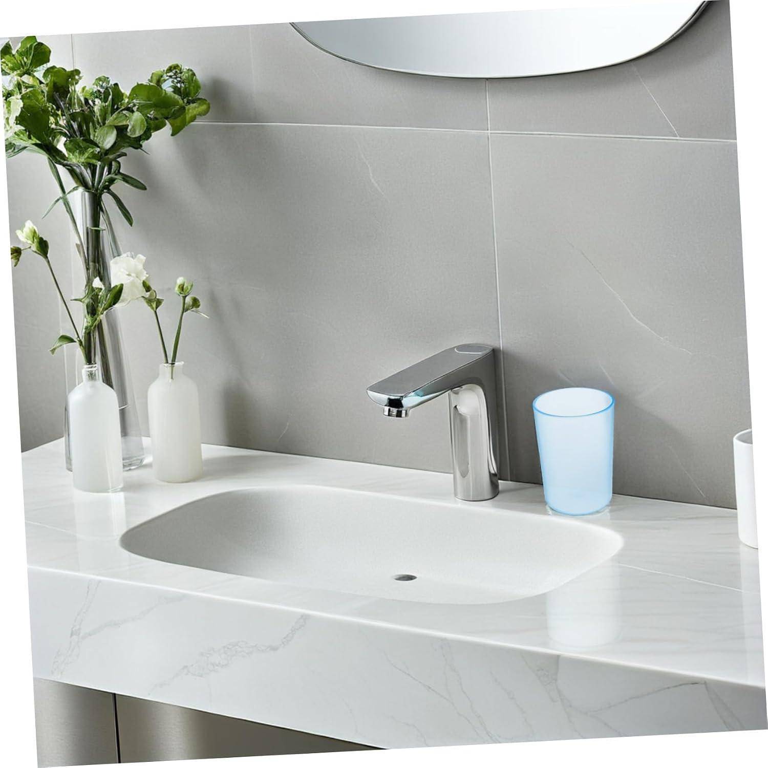 Modernes Badezimmerbecken mit Chromarmatur, Marmortheke, weiße Vasen mit Blumen und ein blauer durchscheinender Becher neben einem Wandspiegel.