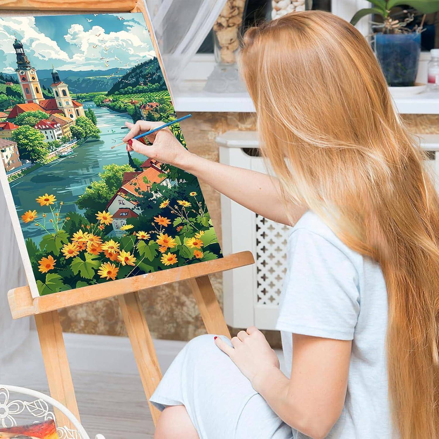 Frau, die eine farbenfrohe Landschaft auf einer Staffelei malt, die einen Fluss, Kirchentürme, Blumen und Häuser in einem hellen Atelier zeigt.