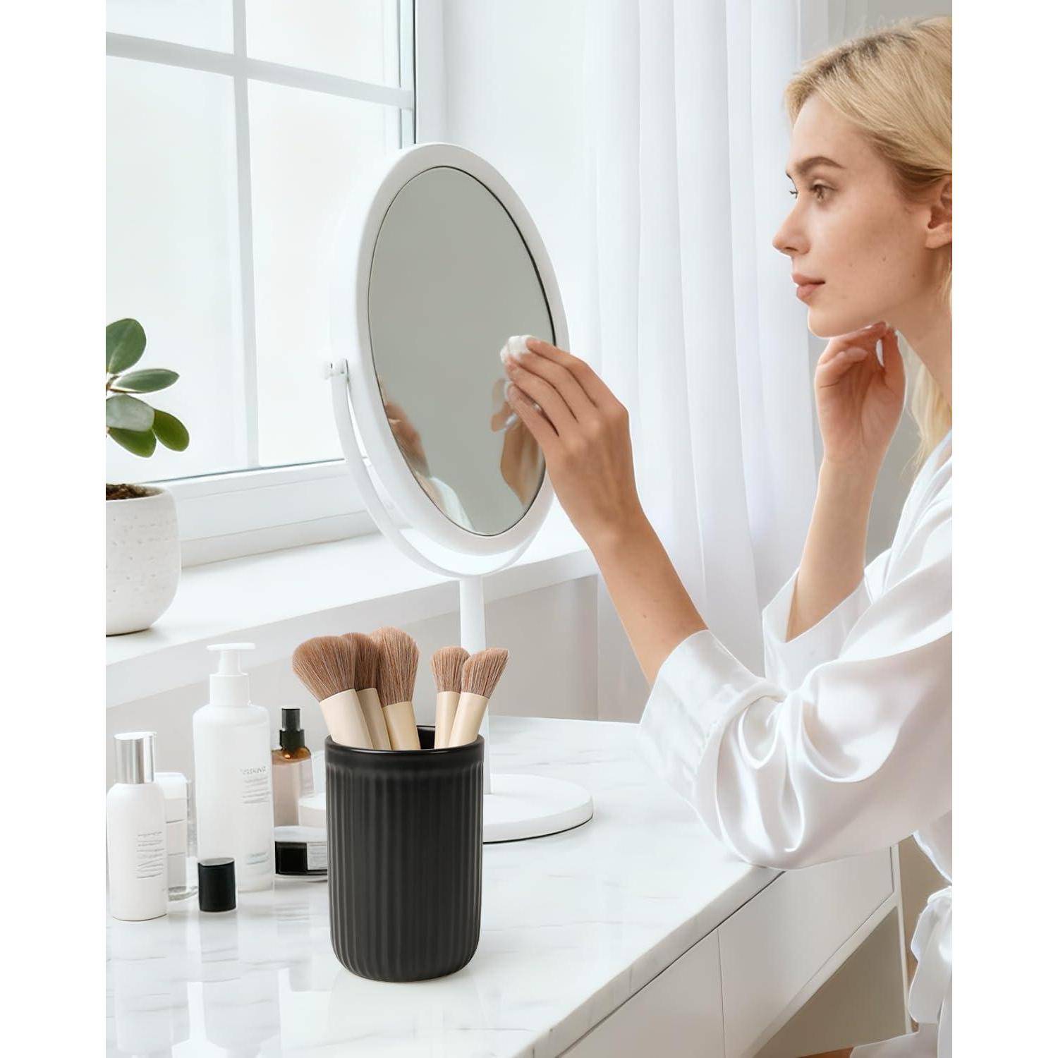 Blonde Frau in einem weißen Bademantel, die sich am Spiegel einer Frisiertischmöbel schminkt, neben Pinseln, Kosmetikprodukten und einer kleinen Pflanze neben einem Fenster.