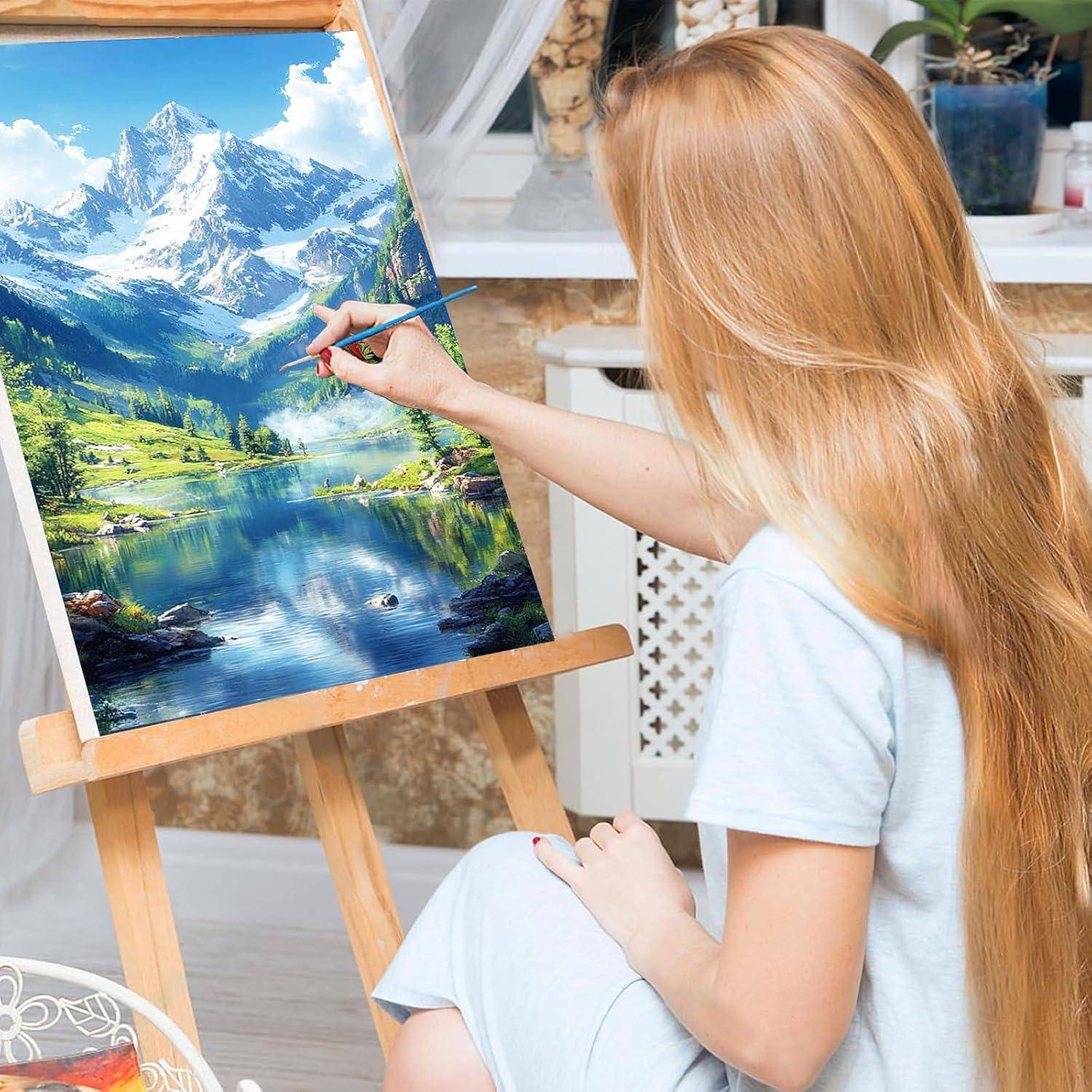 Eine Frau malt eine lebendige Berglandschaft mit See auf einer Staffelei in einem hellen Atelier.