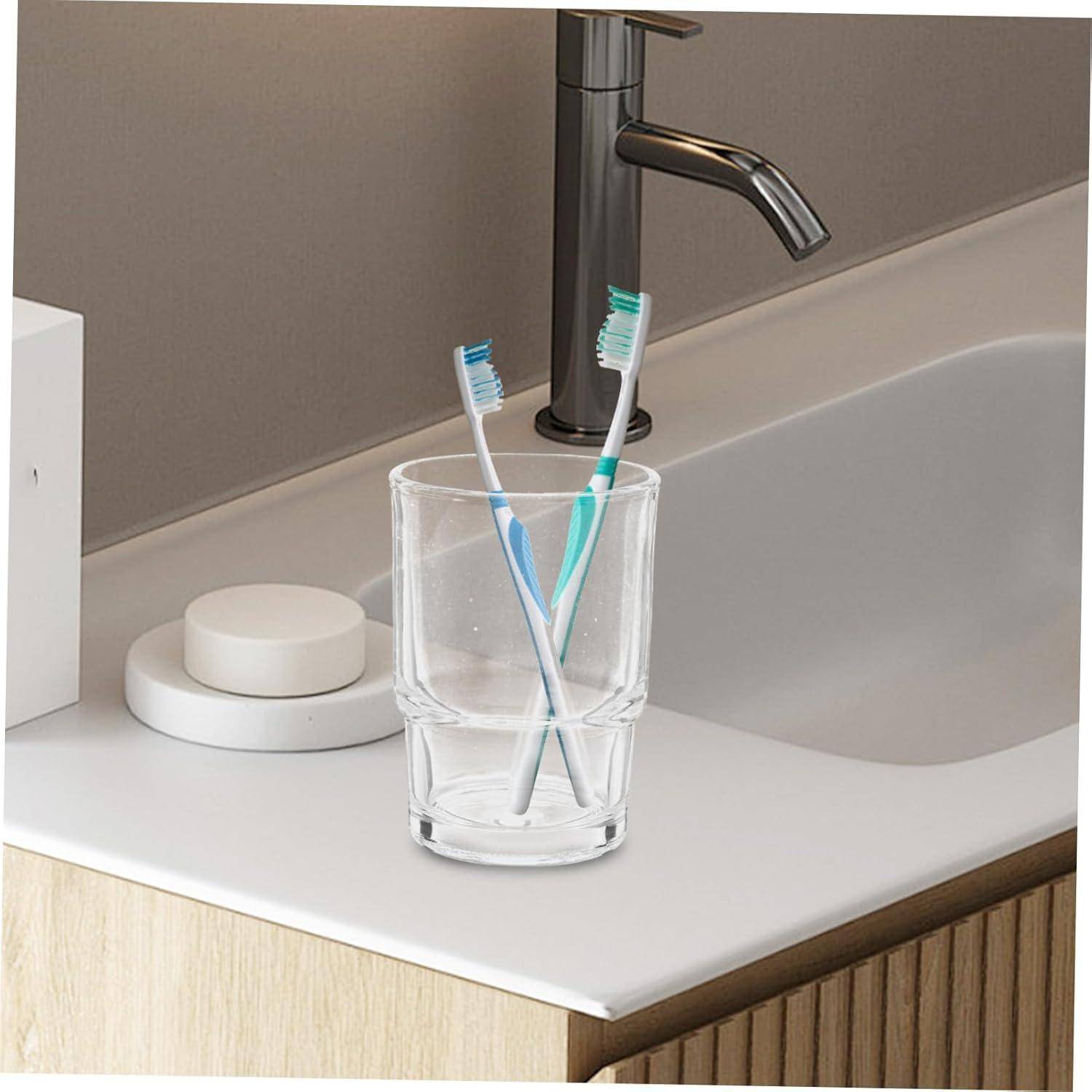 Zwei Zahnbürsten in einem klaren Glas neben einem Badezimmerspülbecken und einem Wasserhahn.