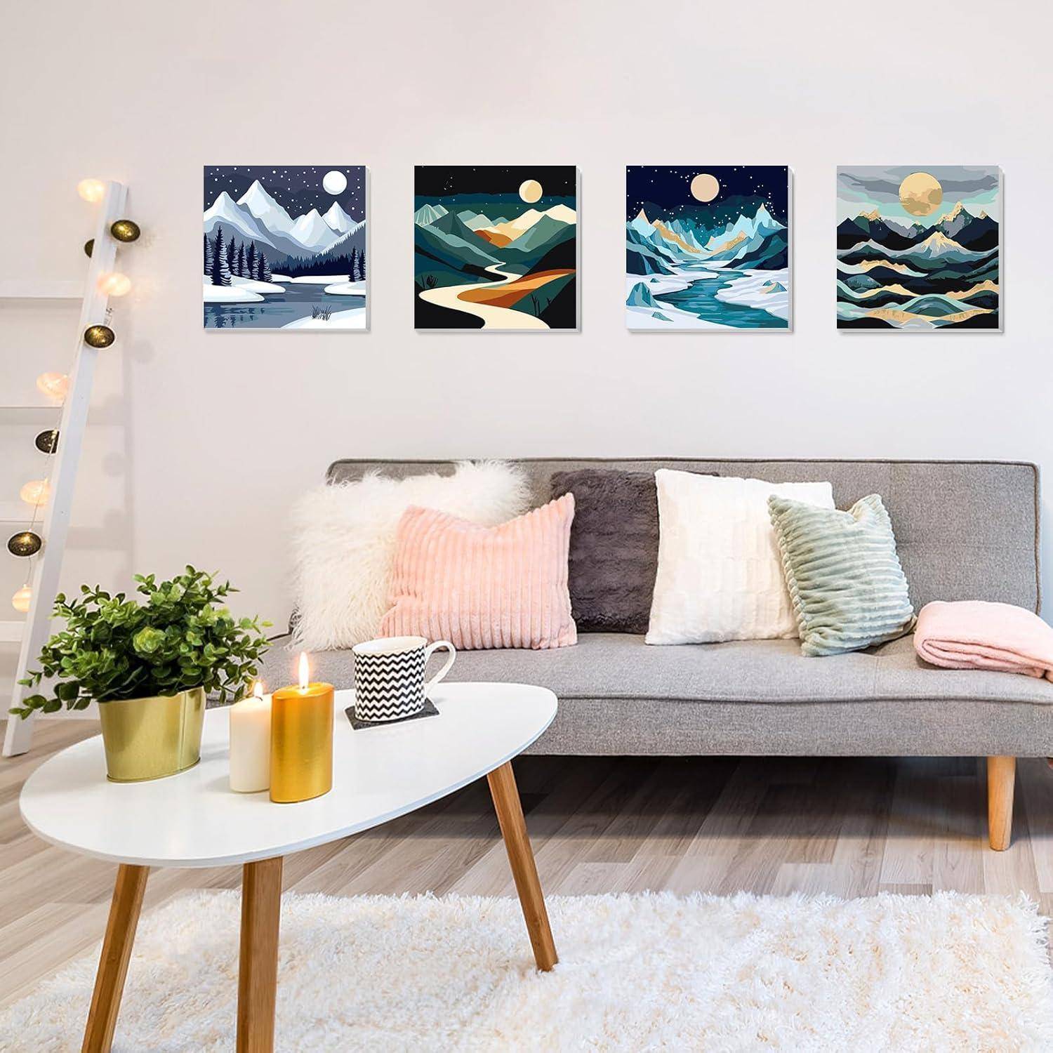 Gemütliches Wohnzimmer mit grauem Sofa, pastellfarbenen Kissen, brennenden Kerzen auf einem weißen Couchtisch, Topfpflanze, Stehleuchte und vier Berglandschaftsdrucken an der Wand.