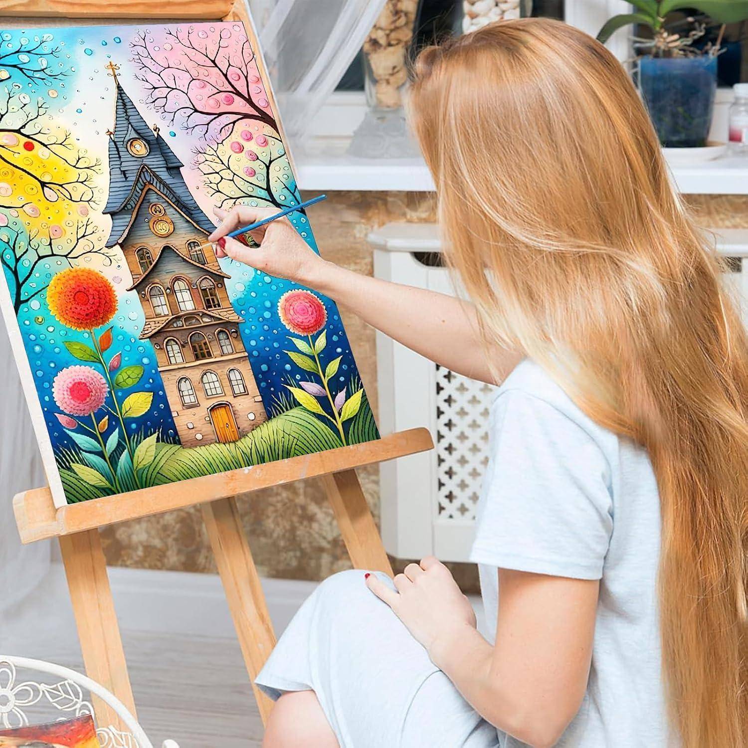 Mädchen malt ein farbenfrohes, verspieltes Haus mit Blumen und einem Baum auf einer Staffelei im Innenraum.