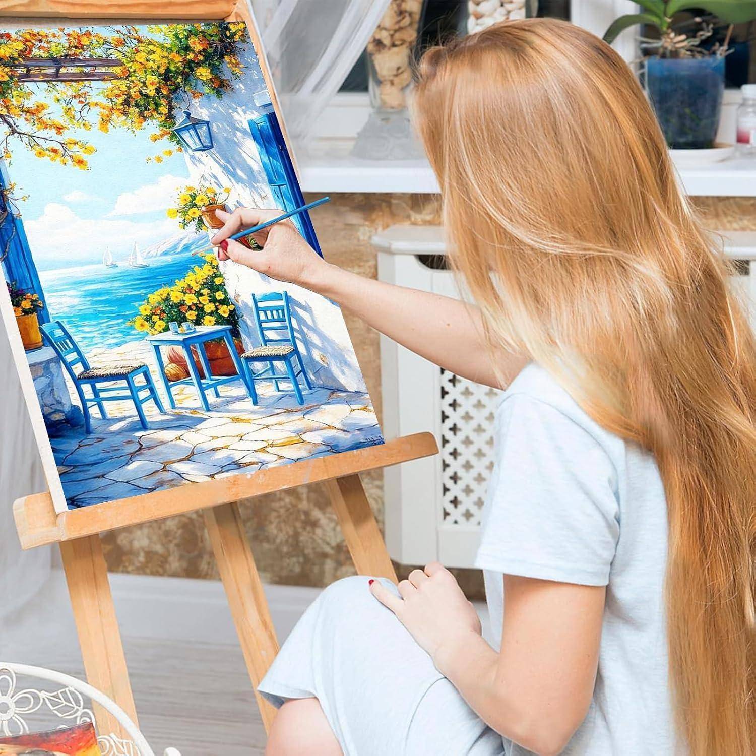 Eine Frau mit langem blondem Haar malt eine helle Strandterrassen-Szene auf einer Leinwand an einer Staffelei.