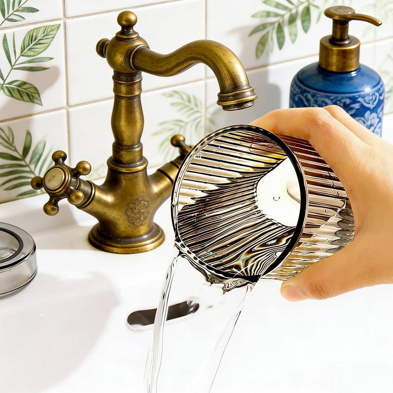 Eine Hand gießt Wasser aus einem Metallbecher in ein weißes Badezimmerbecken neben einem antiken Messinghahn und einem blauen Seifenspender.