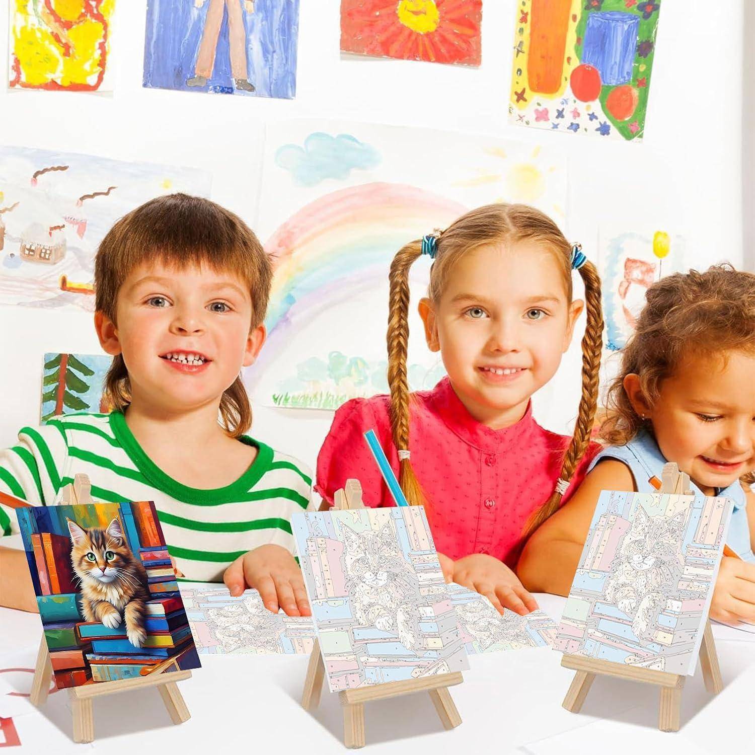 Drei kleine Kinder sitzen an einem Tisch in einem Klassenzimmer, lächeln und bemalen kleine Bildkarten auf Mini-Staffeleien, während bunte Zeichnungen an der Wand hinter ihnen ausgestellt sind.