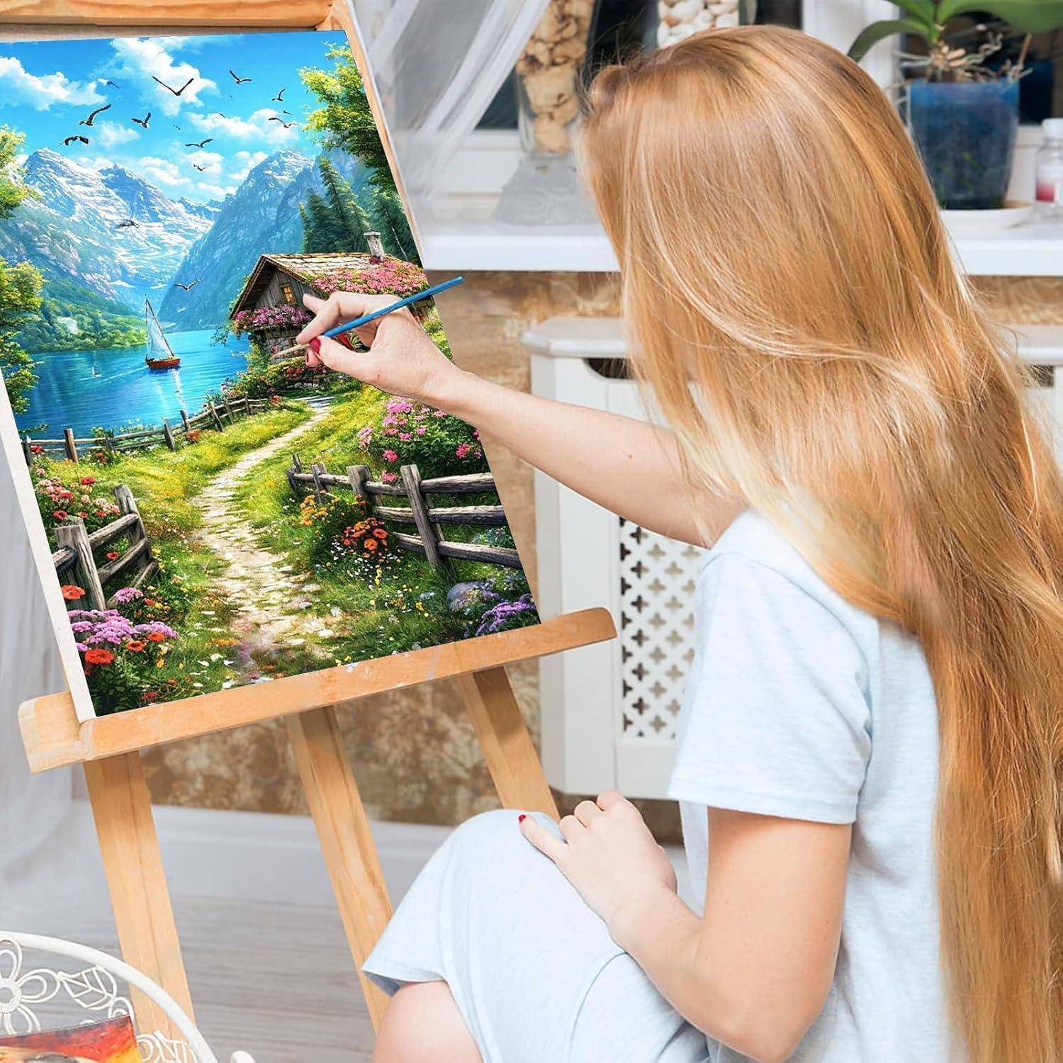 Junge Frau malt eine bunte Landschaft auf einer Staffelei, die einen Bergsee, eine Hütte, einen Pfad und ein Segelboot zeigt.